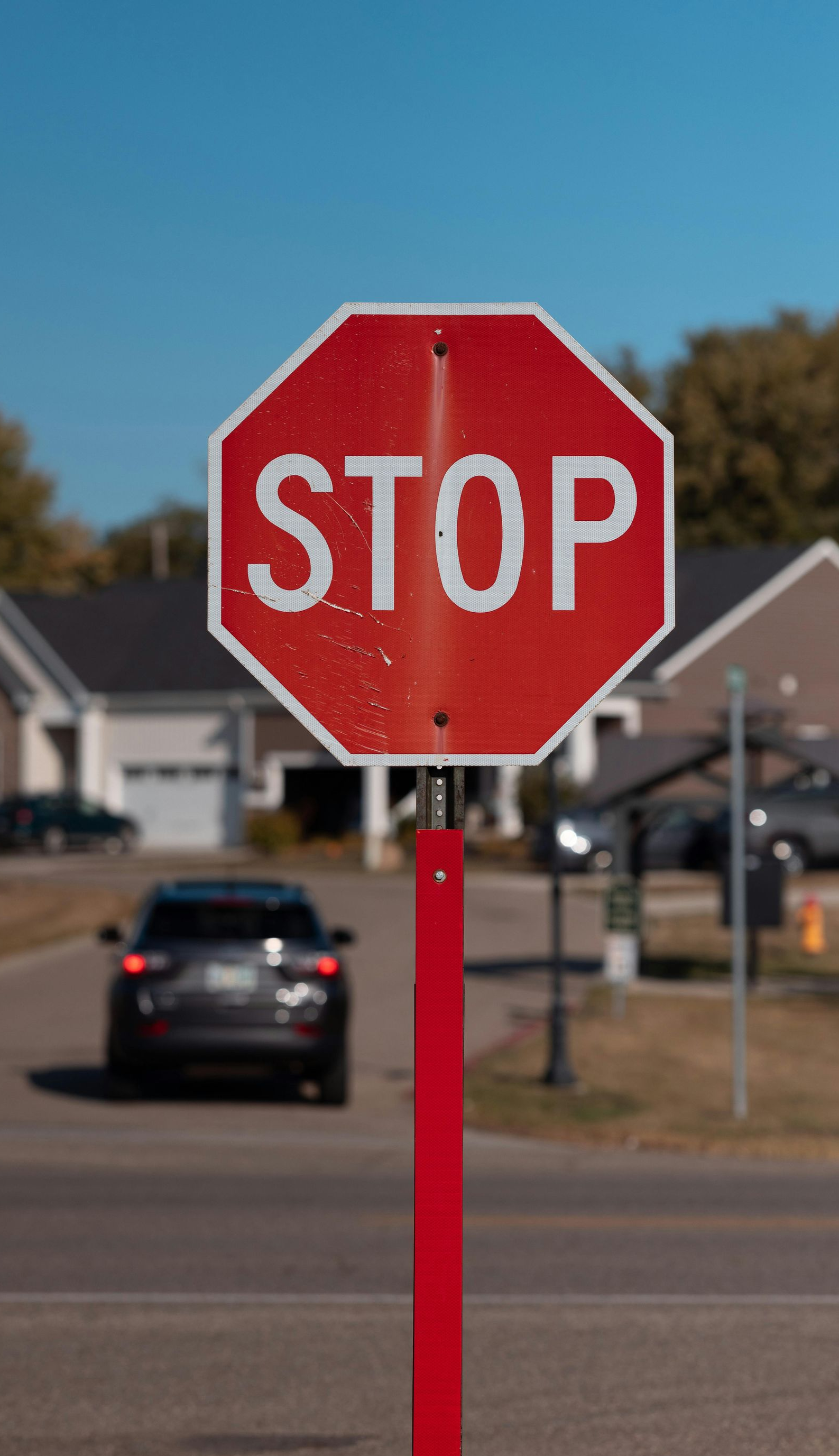 Red stop sign on a red pole in front of a street with a car and houses on a sunny day.