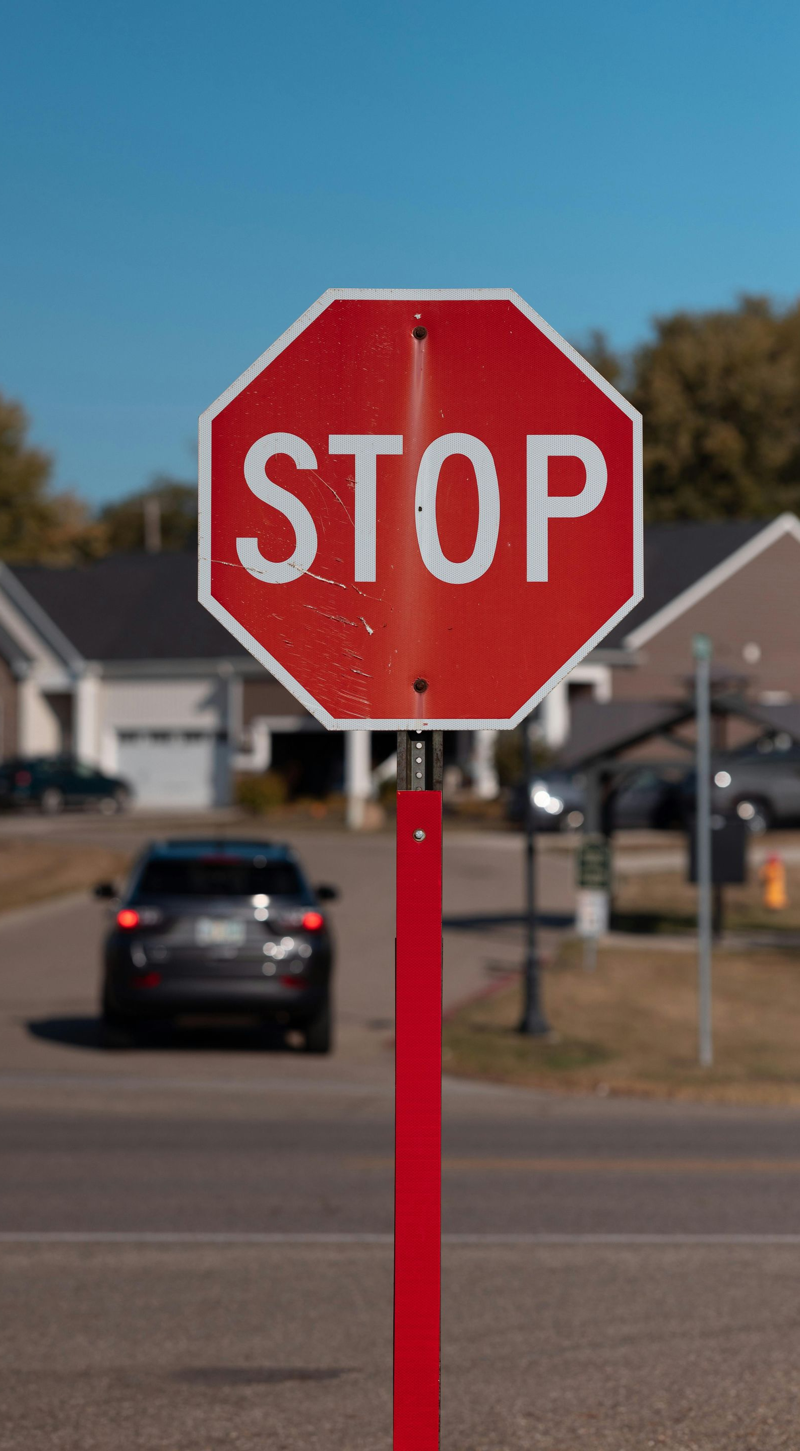 Red stop sign on a red pole in front of a street with a car and houses on a sunny day.