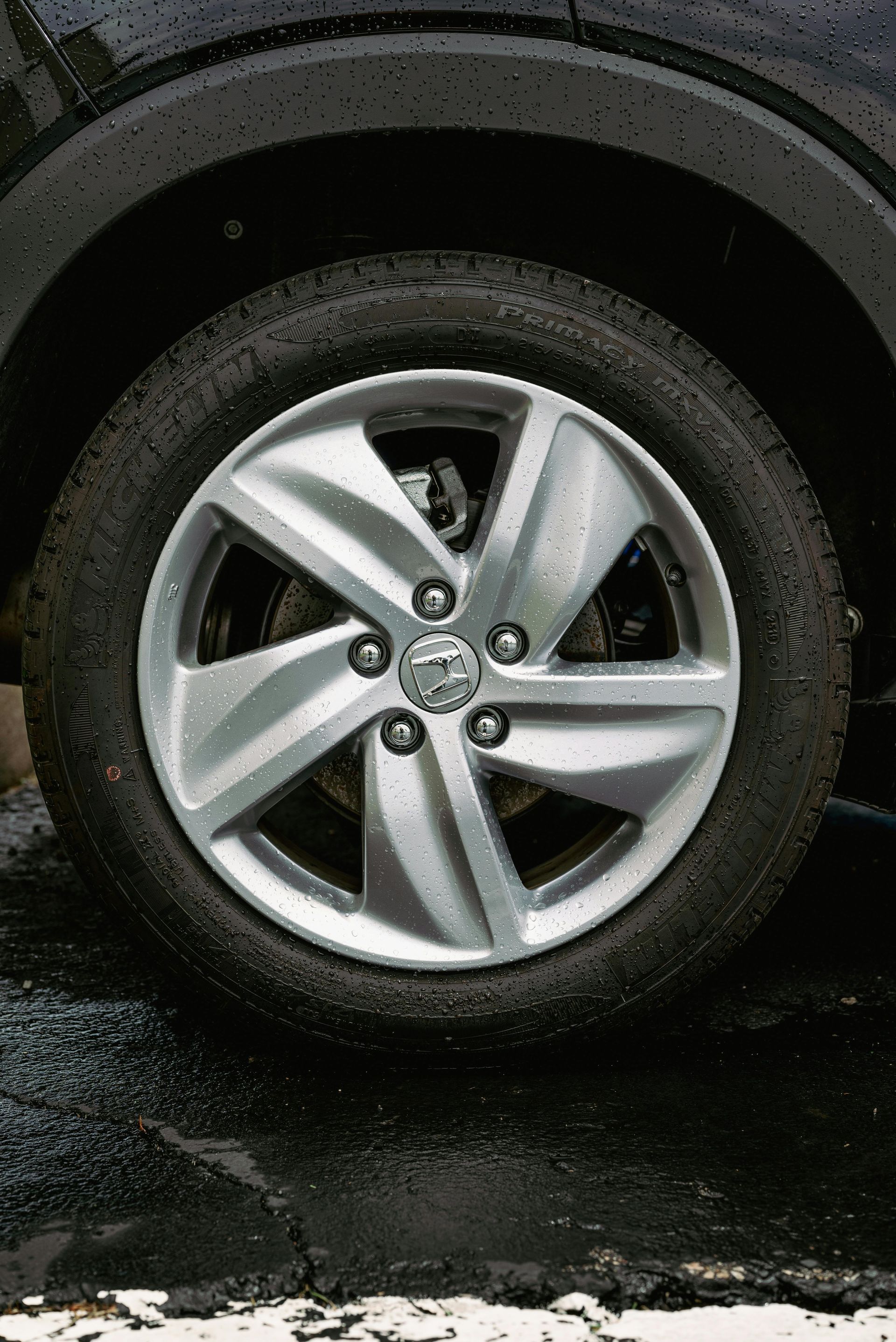 A close-up view of a five-spoke silver alloy car wheel with a circular logo in the center, wet from rain.
