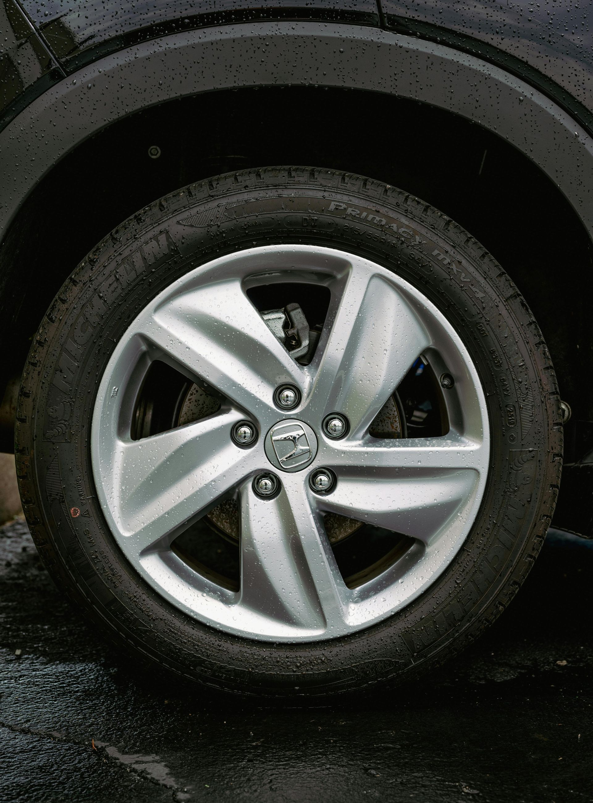 A close-up view of a five-spoke silver alloy car wheel with a circular logo in the center, wet from rain.