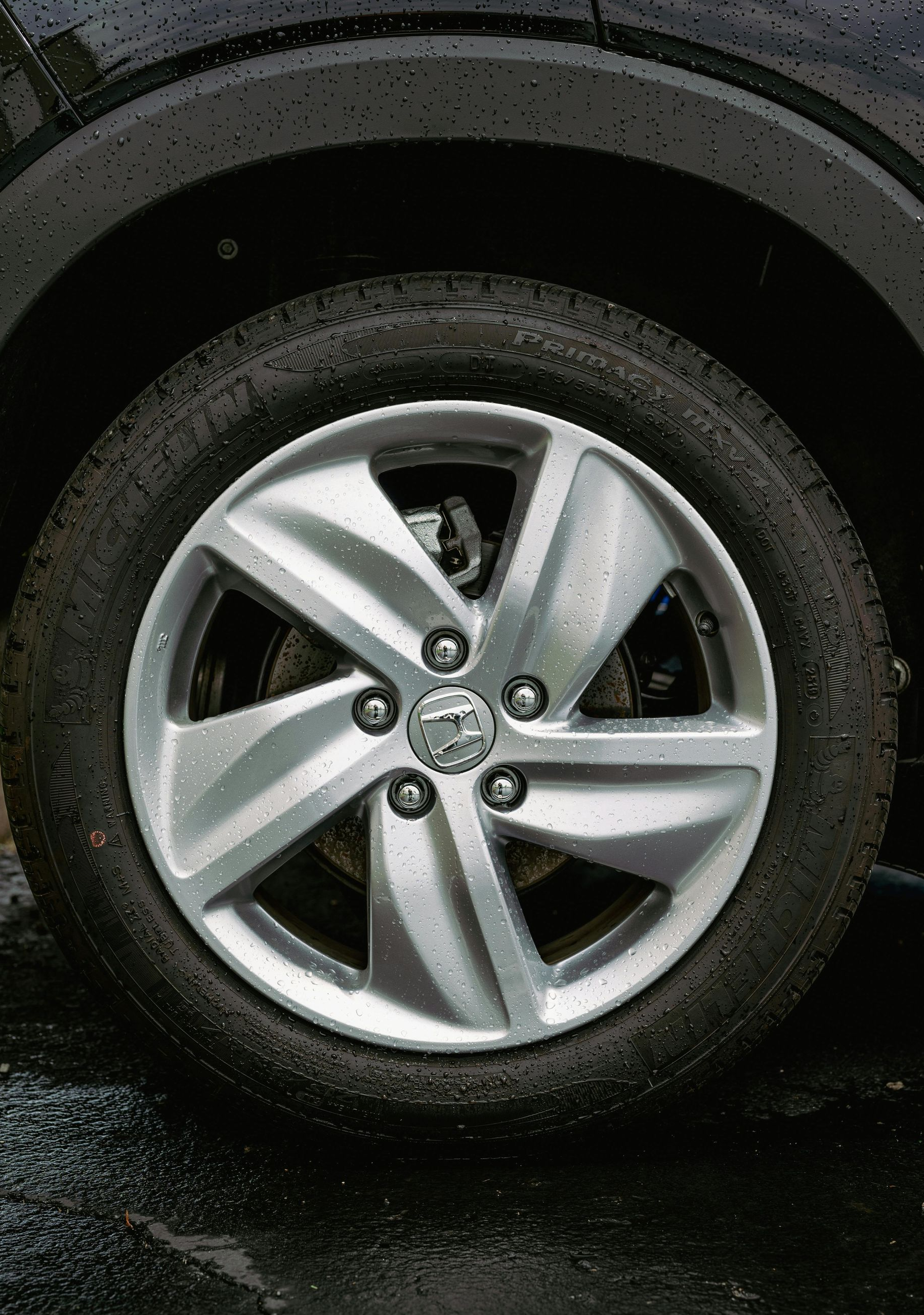 A close-up view of a five-spoke silver alloy car wheel with a circular logo in the center, wet from rain.