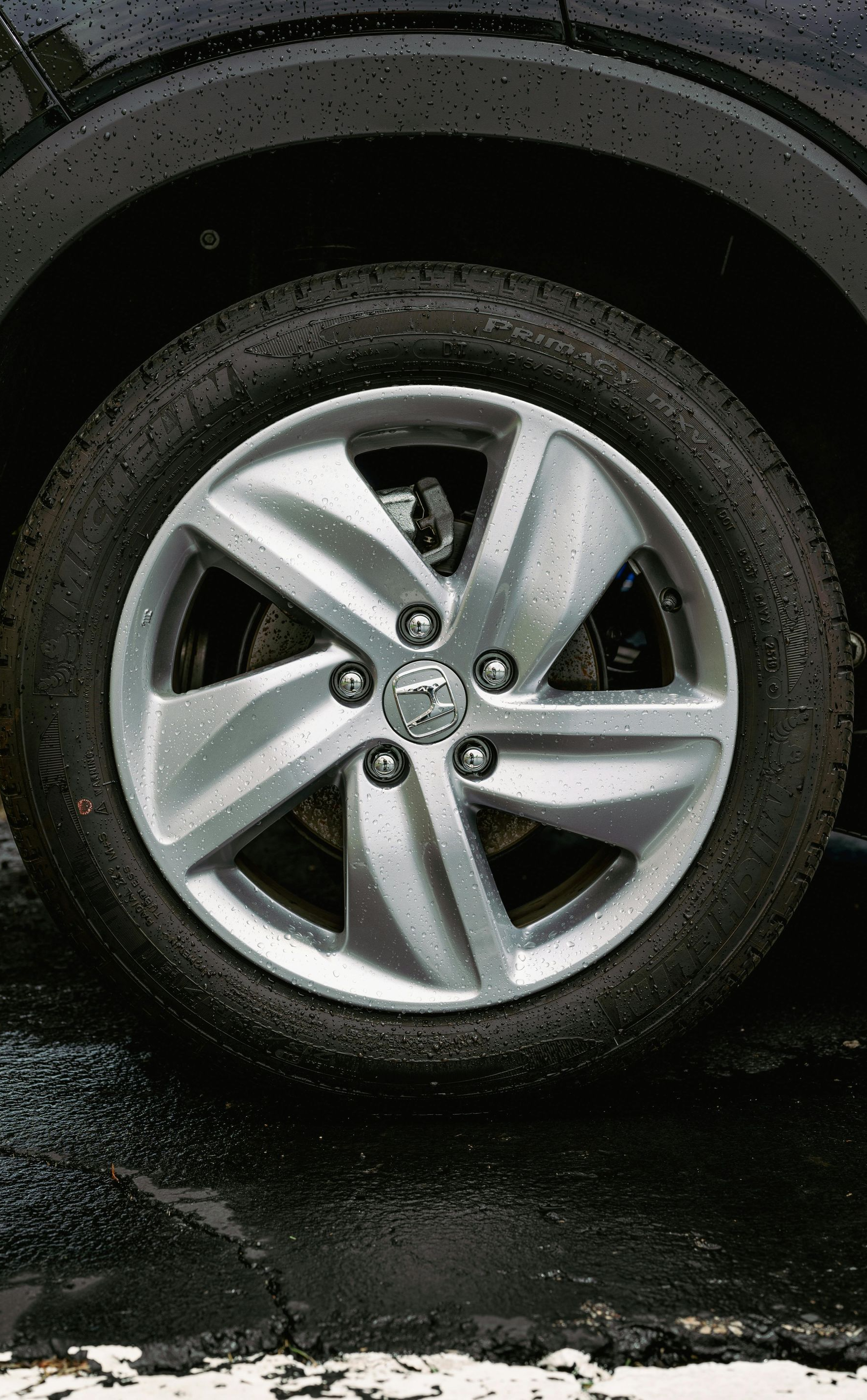 A close-up view of a five-spoke silver alloy car wheel with a circular logo in the center, wet from rain.