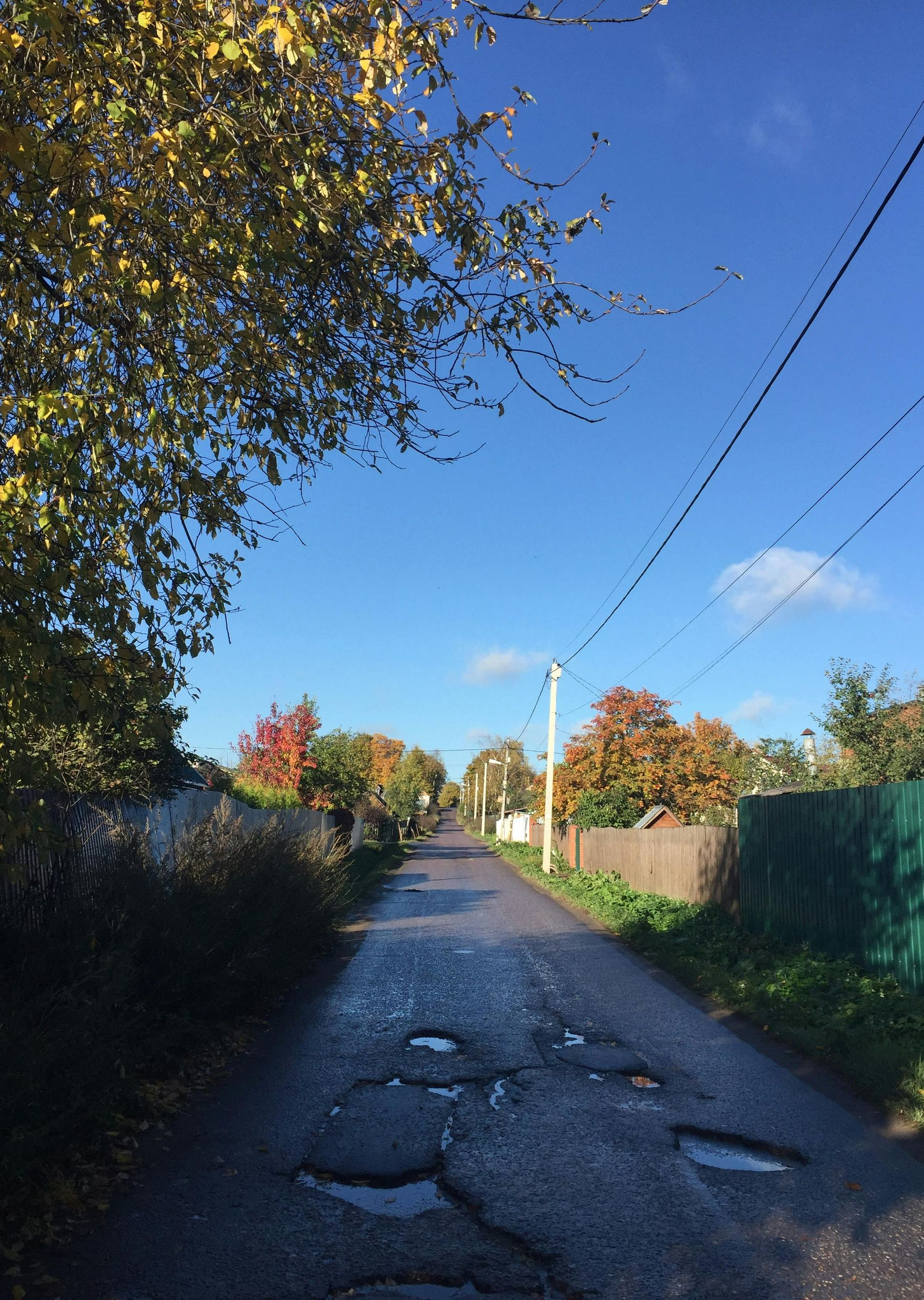 Paved road with potholes in a rural area, lined with houses, trees with autumn colors, under a blue sky.