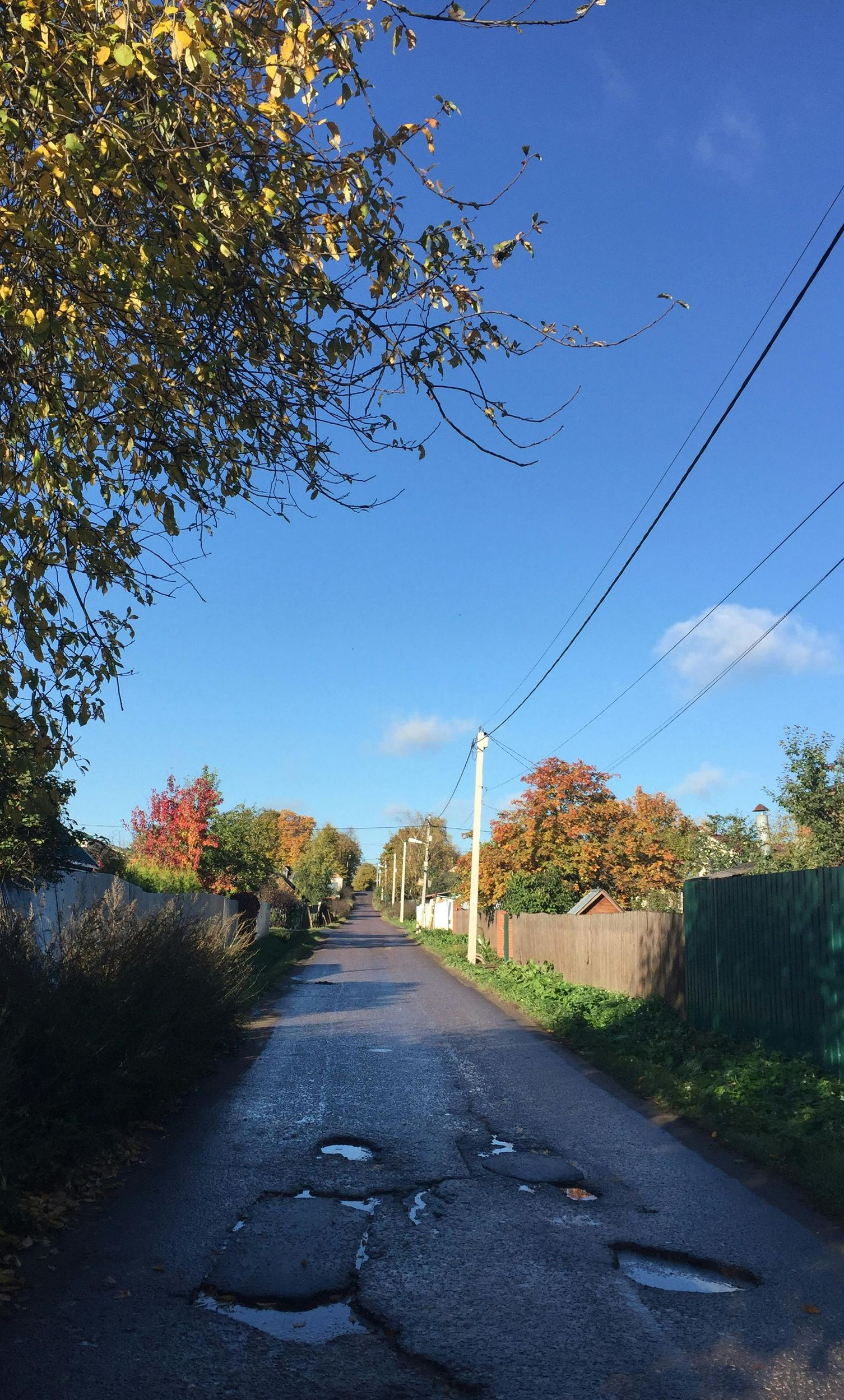 Paved road with potholes in a rural area, lined with houses, trees with autumn colors, under a blue sky.