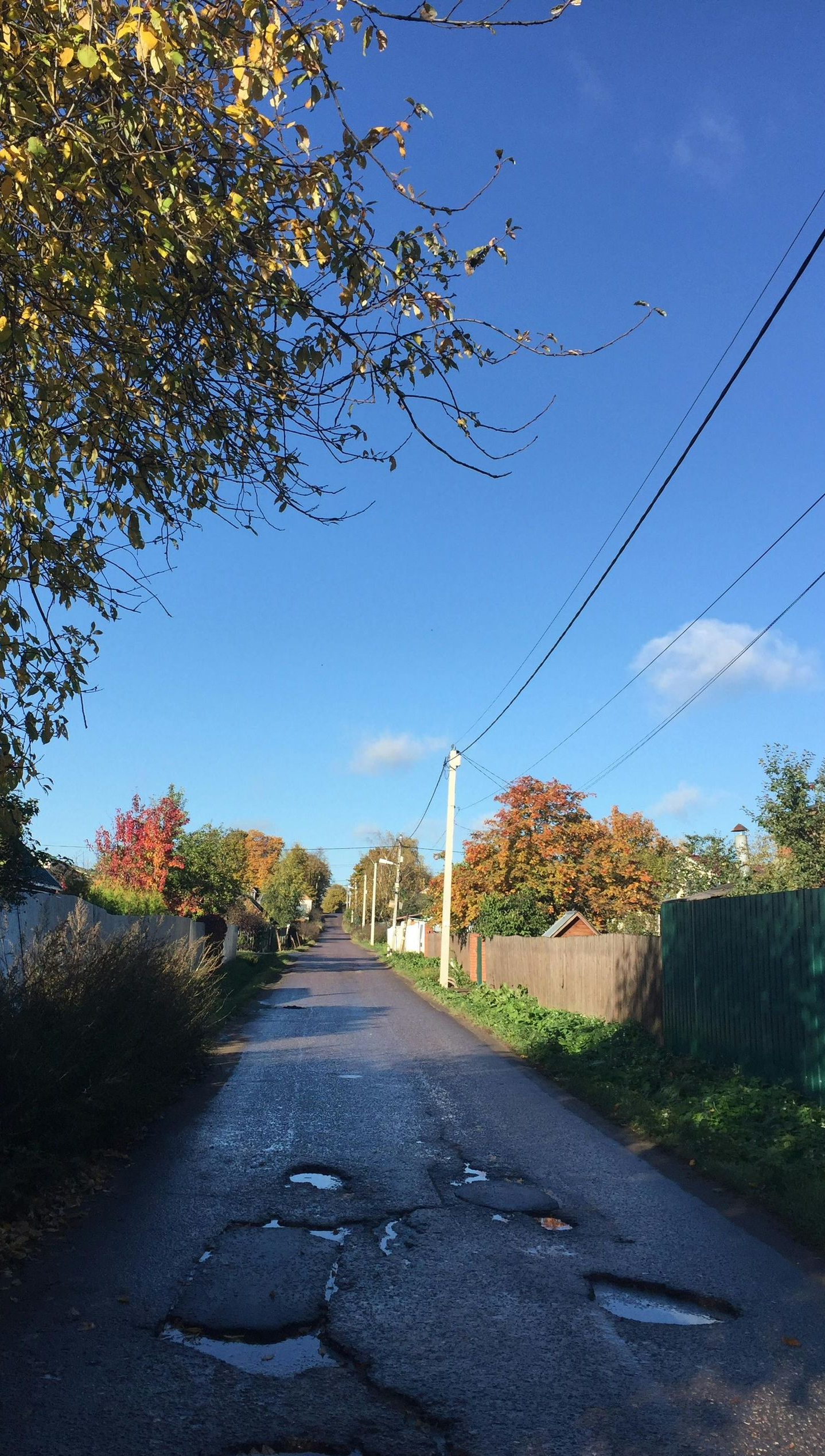 Paved road with potholes in a rural area, lined with houses, trees with autumn colors, under a blue sky.