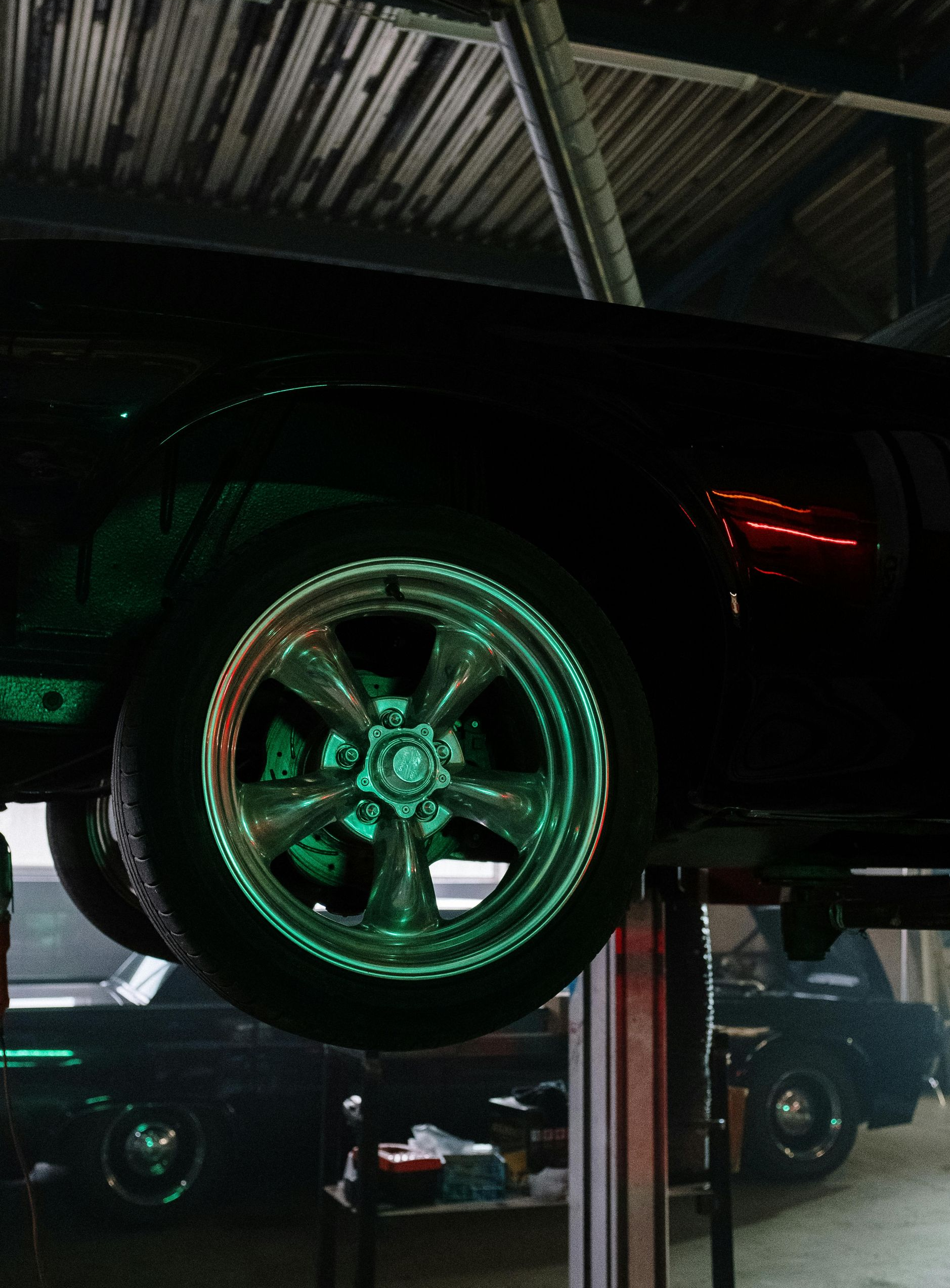 Black classic car raised on a lift in a garage, rims highlighted by green light.