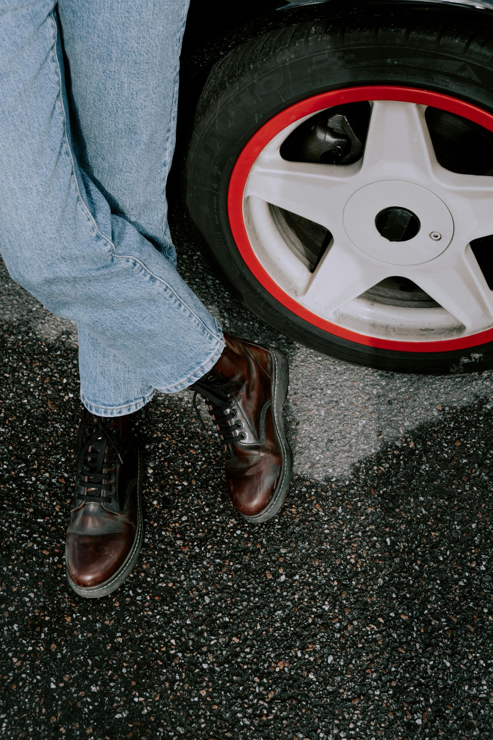 Person in jeans and brown boots leaning against a car wheel with a red rim on asphalt.