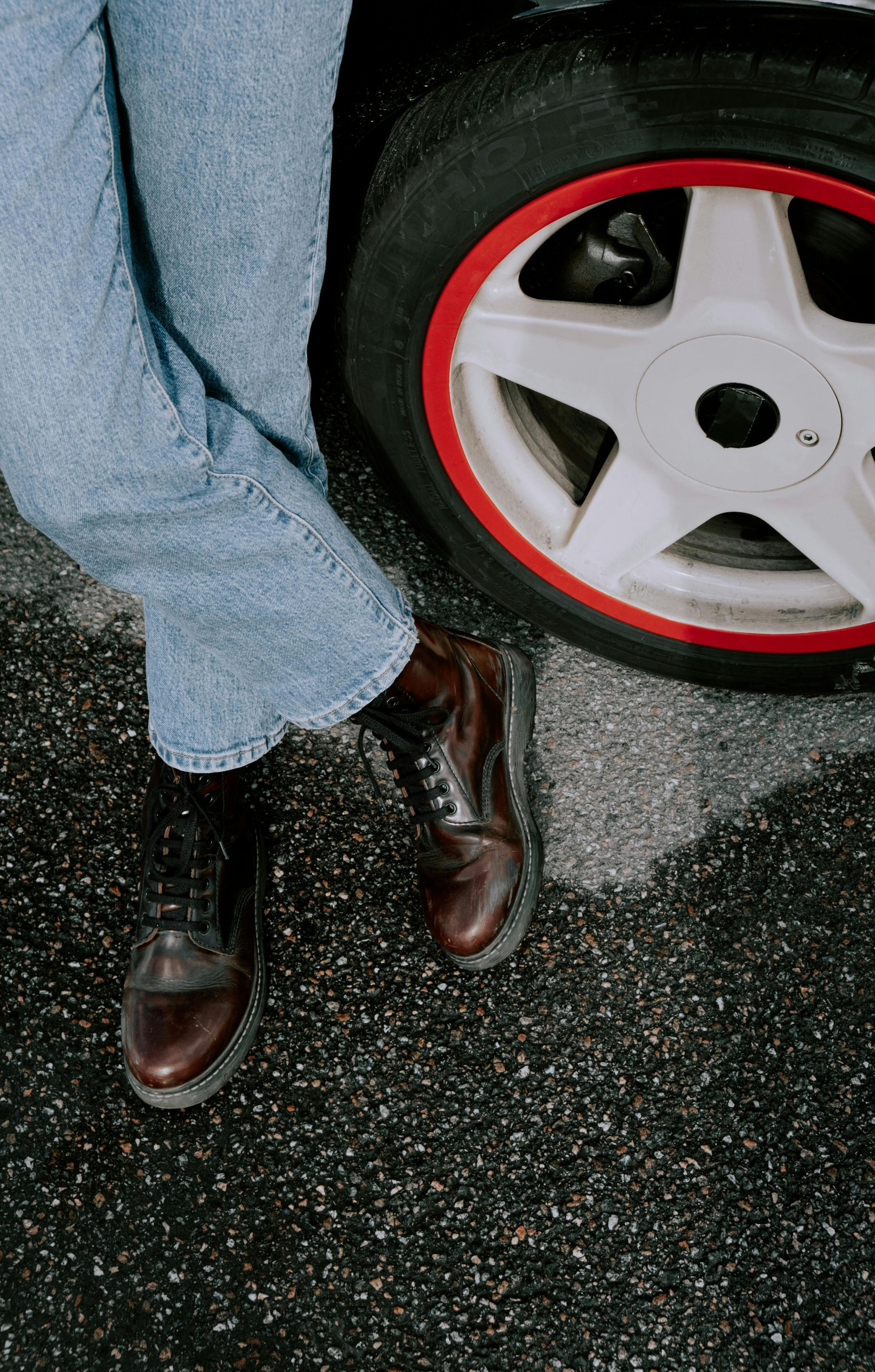 Person in jeans and brown boots leaning against a car wheel with a red rim on asphalt.