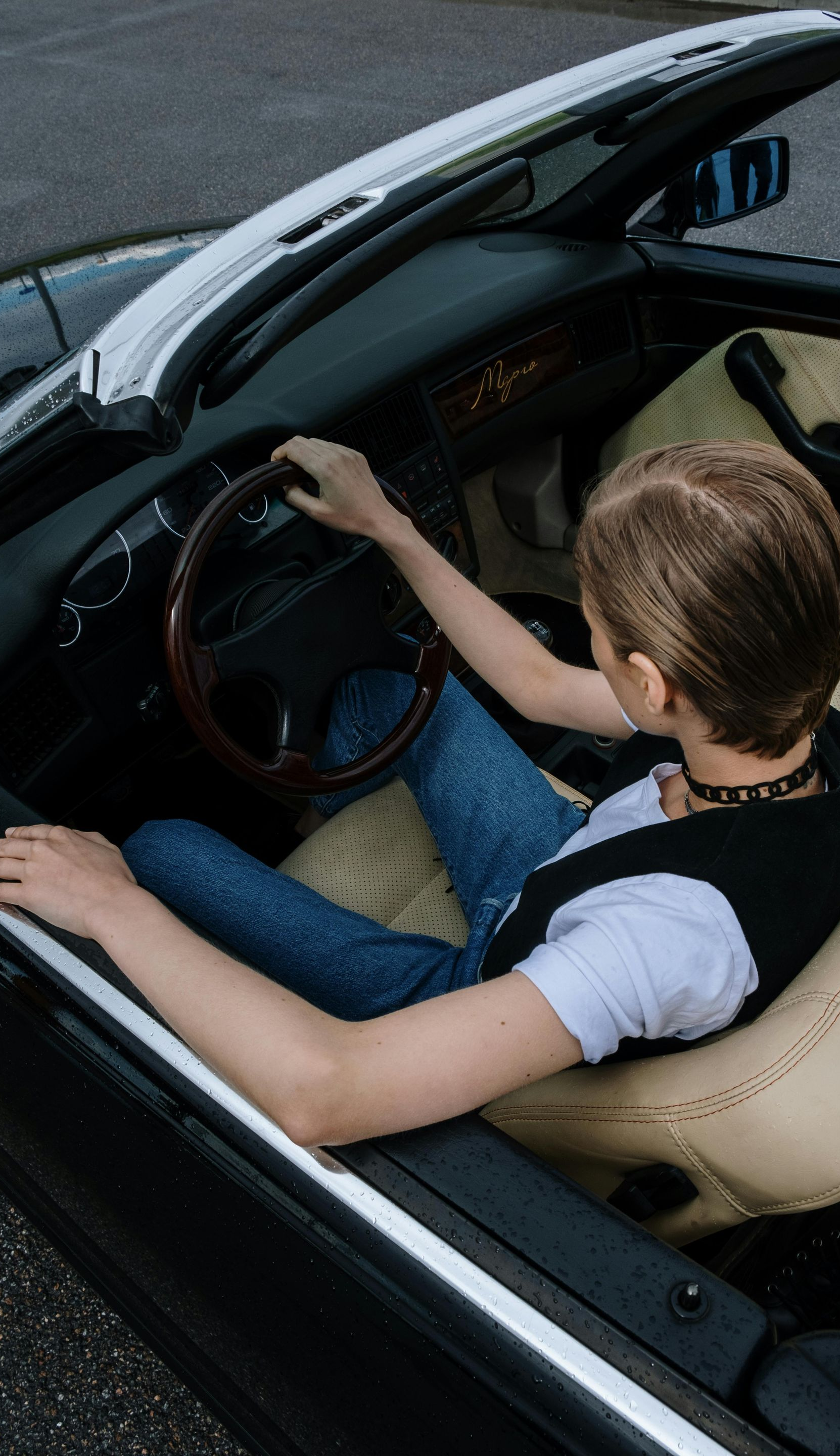 Person sits in a black convertible, holding the steering wheel. They wear a black vest and jeans.