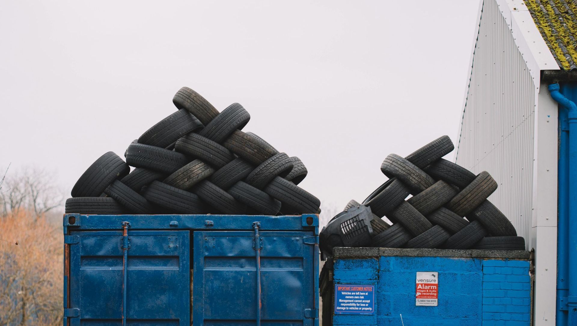 Two blue containers overflowing with stacks of old tires, next to a white and blue building under a gray sky.