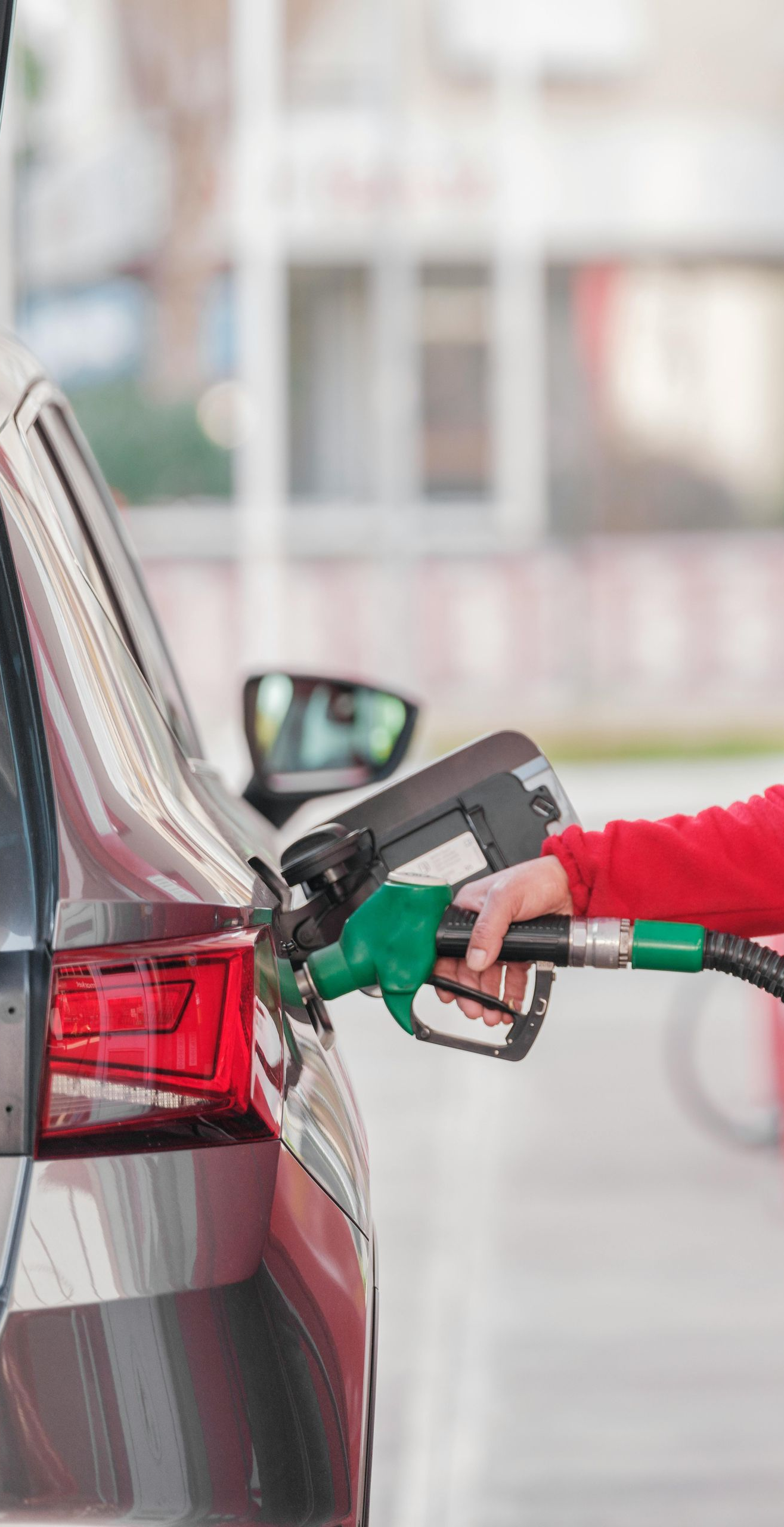 Car being refueled at a gas station; a person's hand holds the green nozzle, filling the fuel tank.