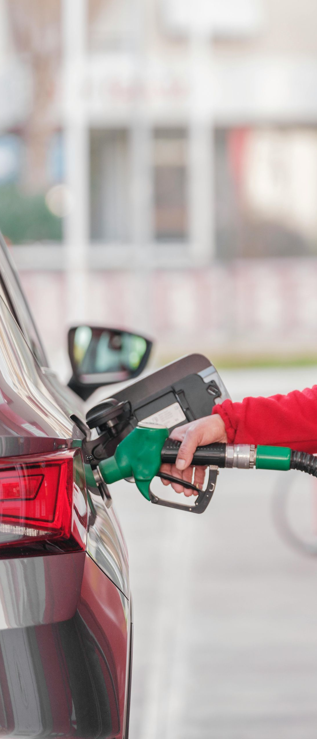 Car being refueled at a gas station; a person's hand holds the green nozzle, filling the fuel tank.