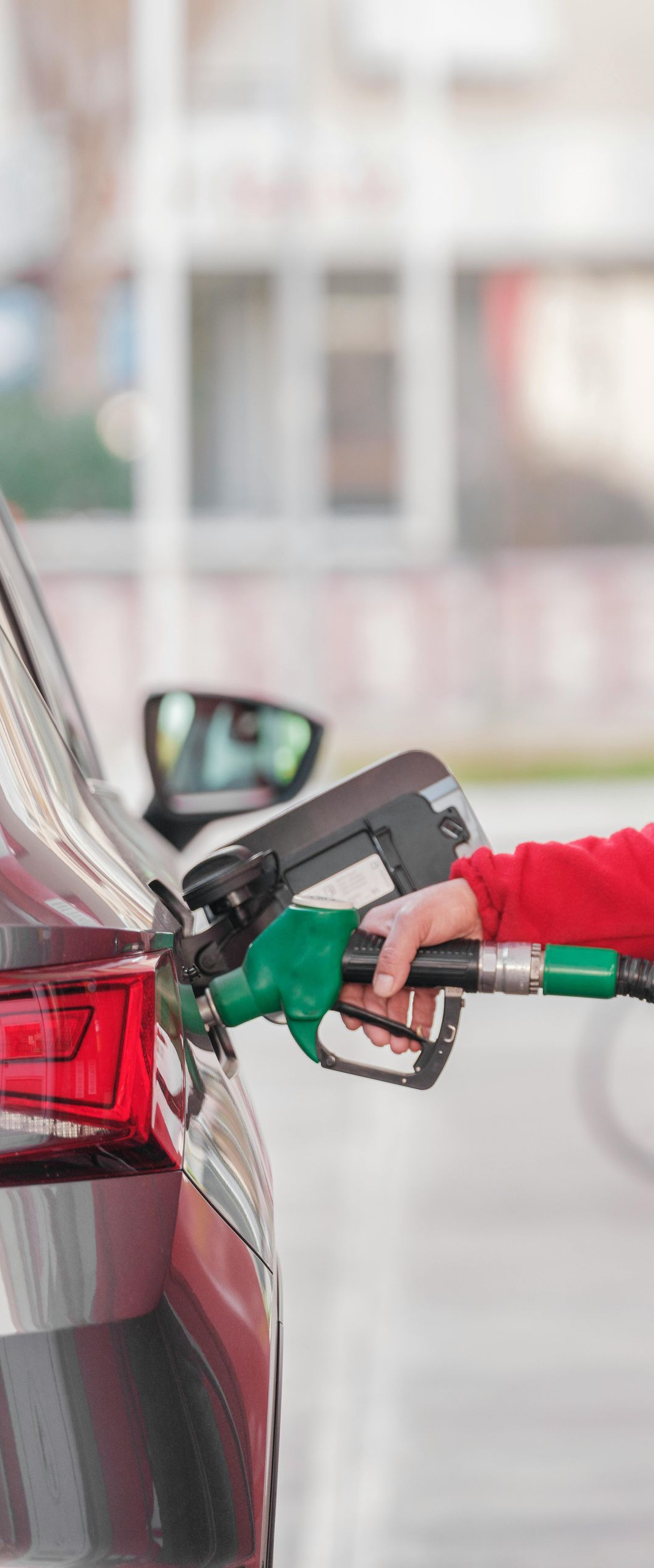 Car being refueled at a gas station; a person's hand holds the green nozzle, filling the fuel tank.