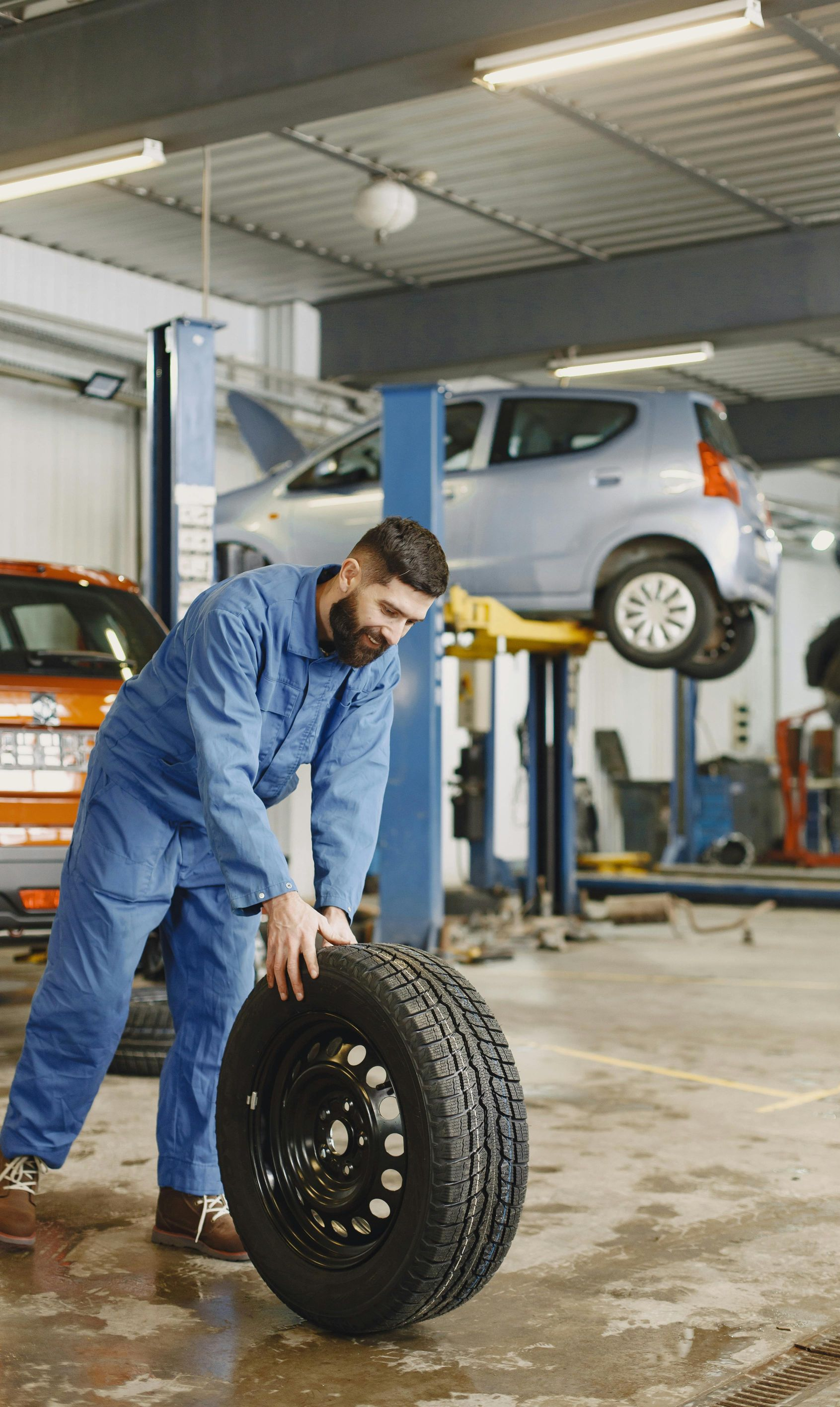 Mechanic rolling a tire in a garage, another car on a lift in the background.