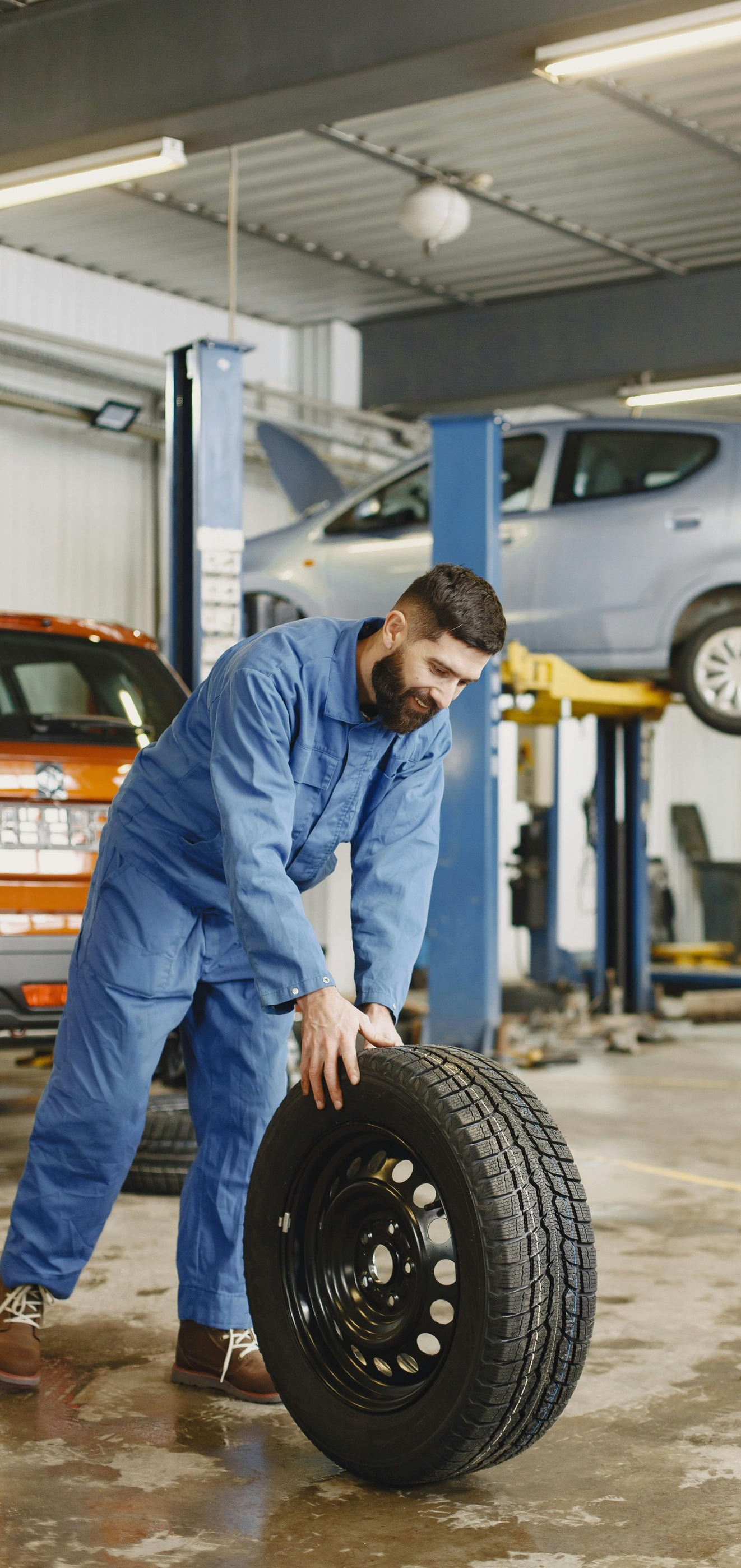 Mechanic rolling a tire in a garage, another car on a lift in the background.