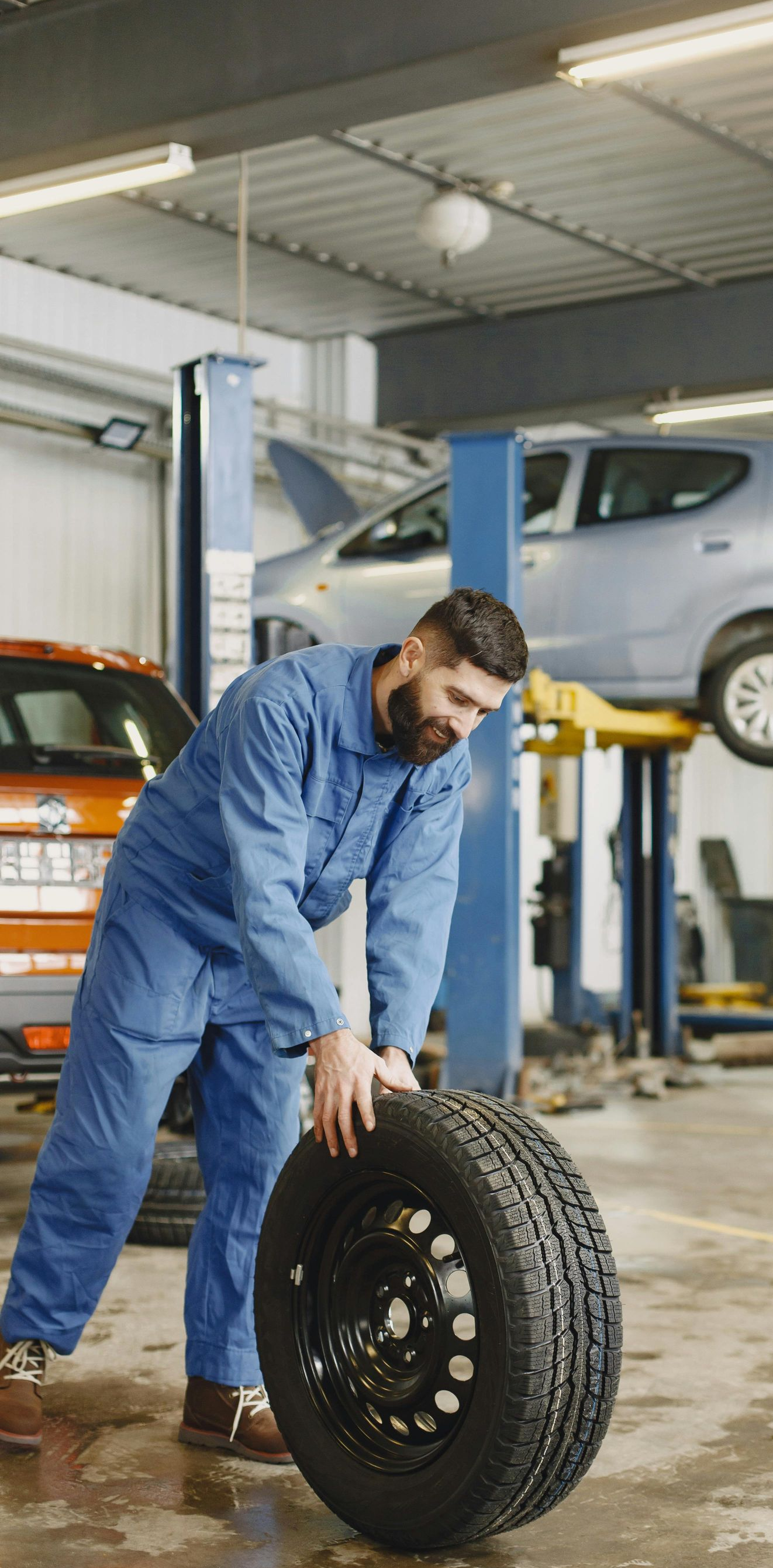 Mechanic rolling a tire in a garage, another car on a lift in the background.