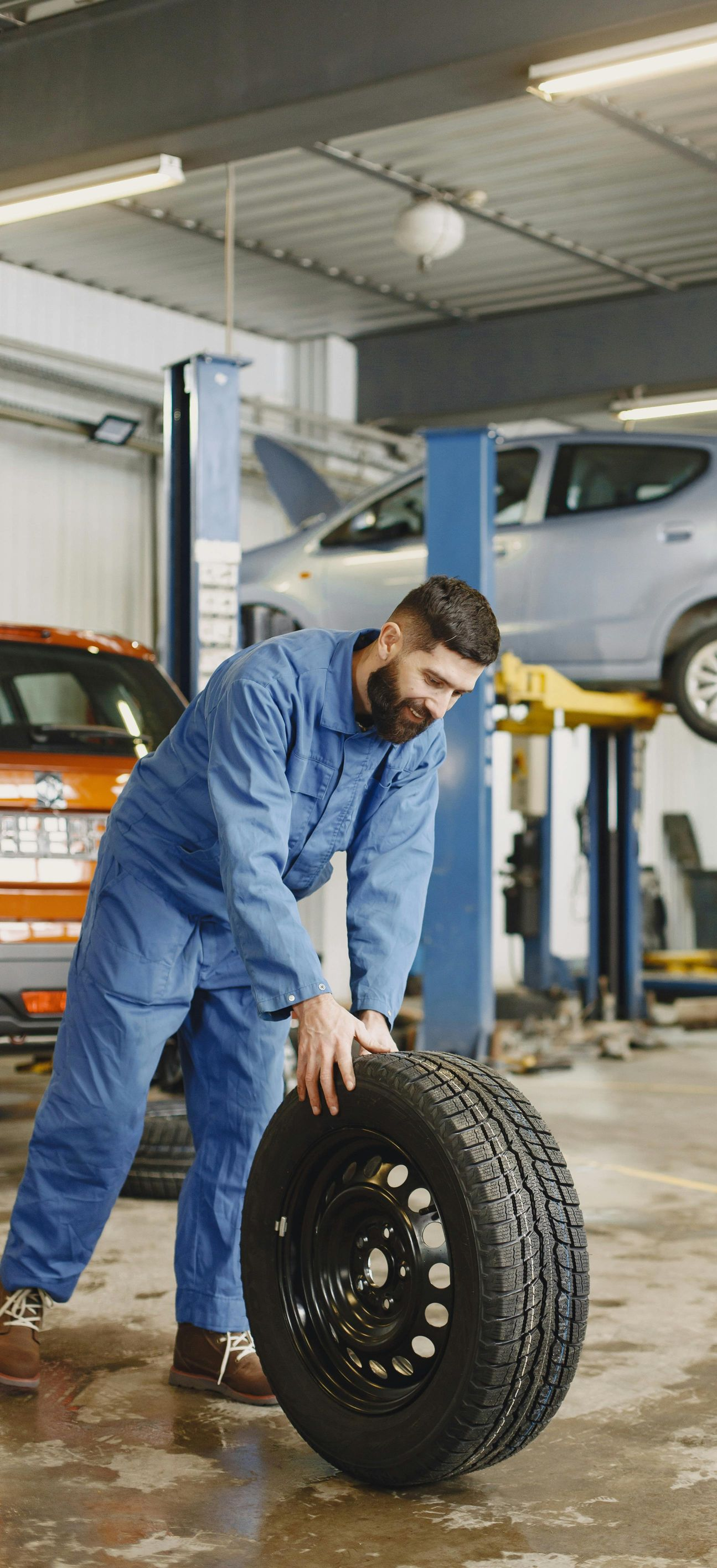 Mechanic rolling a tire in a garage, another car on a lift in the background.