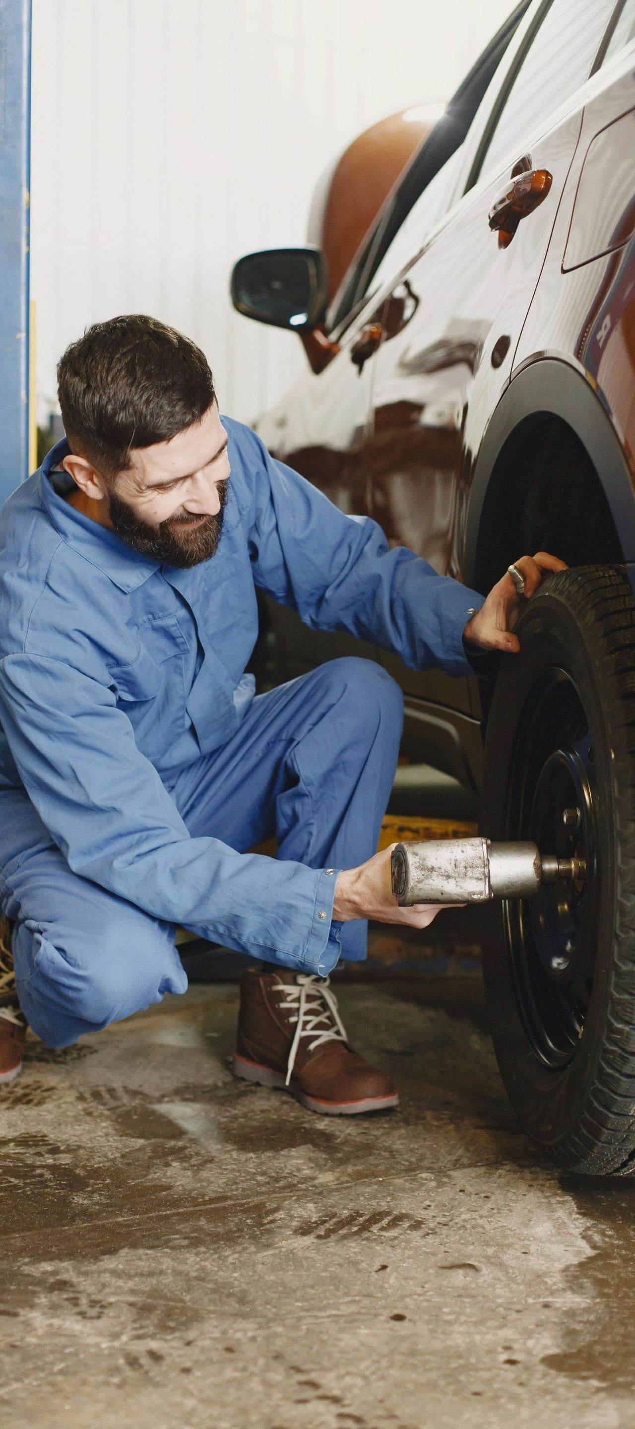 A mechanic in blue overalls kneels, using a tool to work on a car tire in a garage. He has a beard and focused expression.