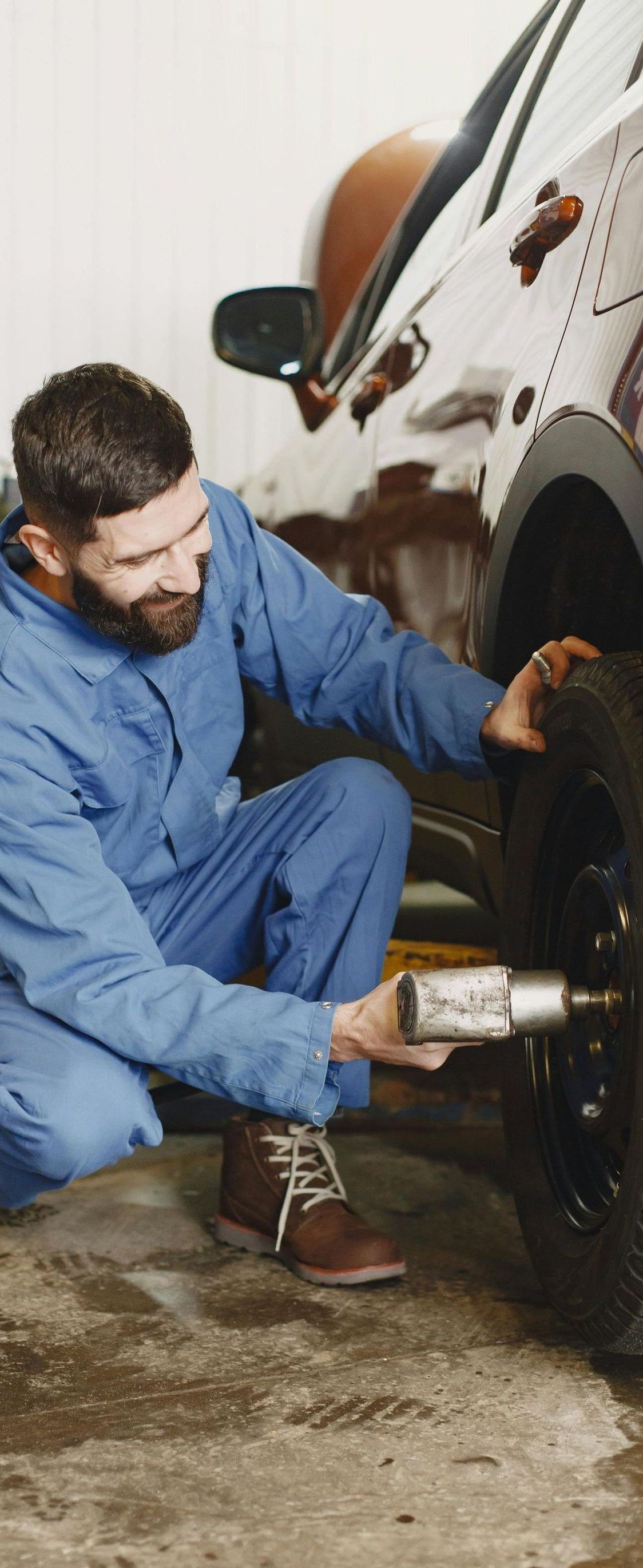 A mechanic in blue overalls kneels, using a tool to work on a car tire in a garage. He has a beard and focused expression.
