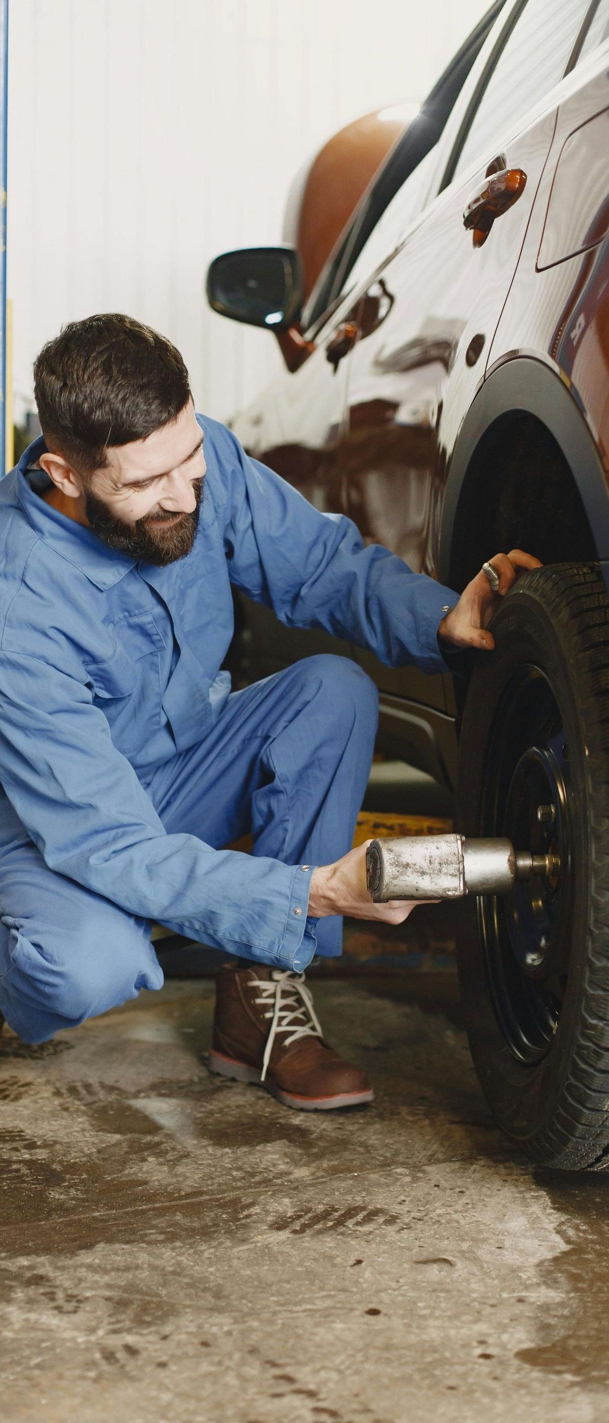 A mechanic in blue overalls kneels, using a tool to work on a car tire in a garage. He has a beard and focused expression.