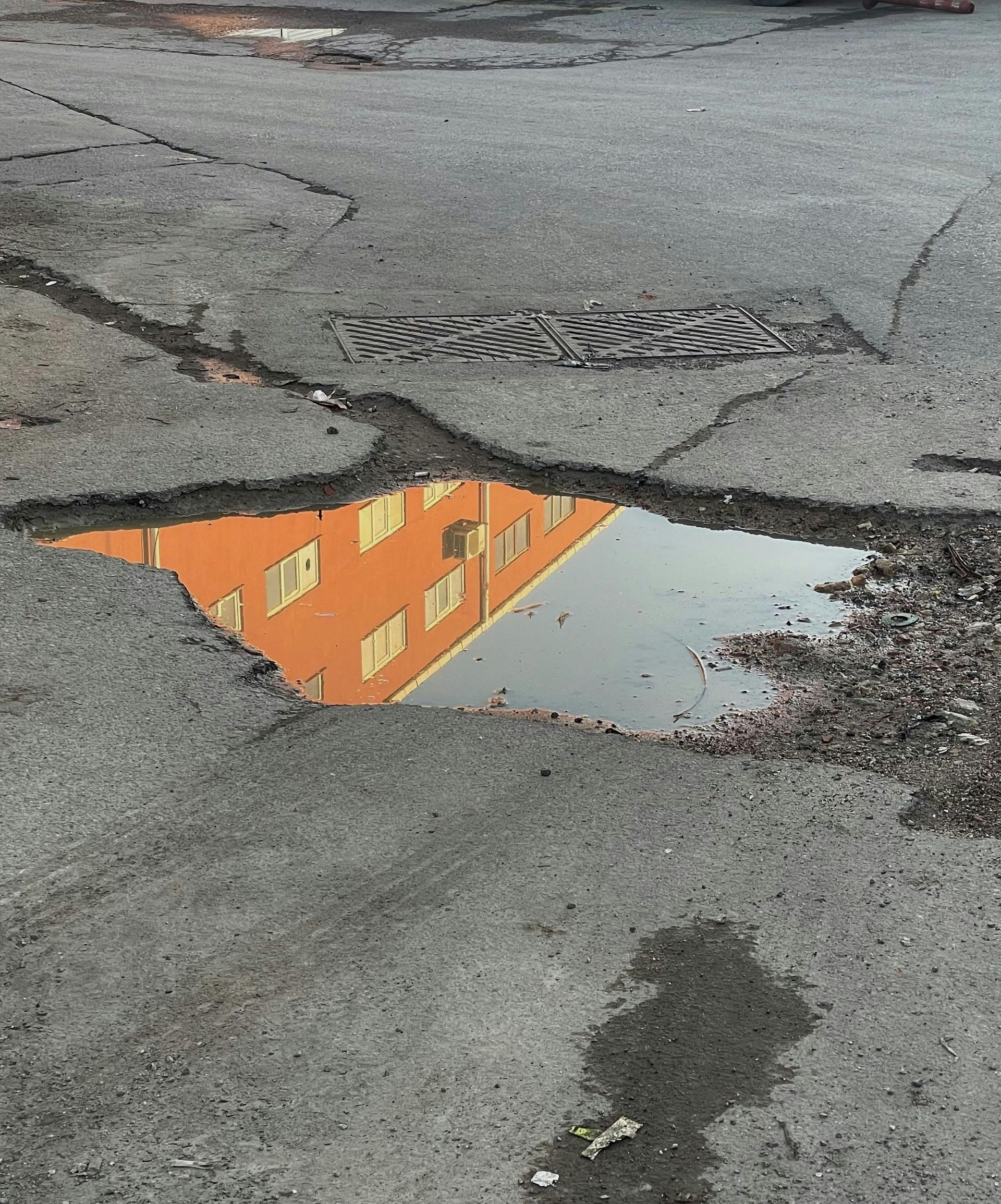 A puddle in a large pothole on a cracked asphalt road reflects a multi-story orange building.