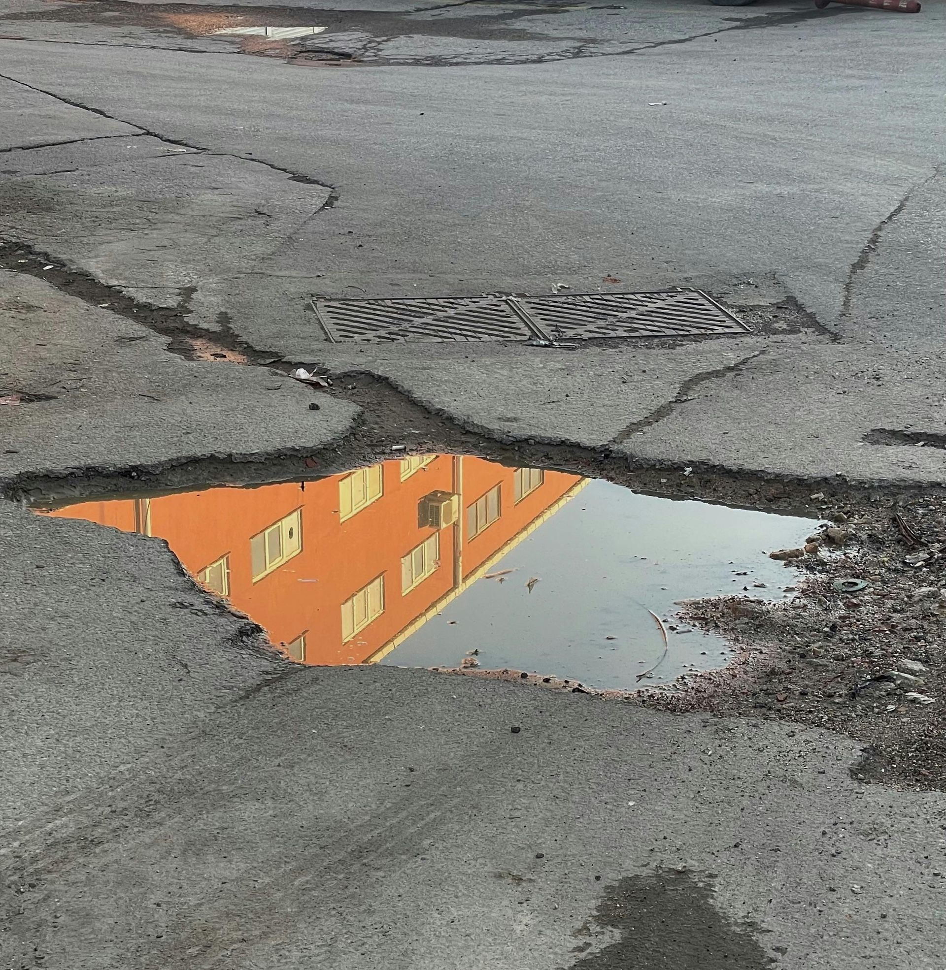 A puddle in a large pothole on a cracked asphalt road reflects a multi-story orange building.