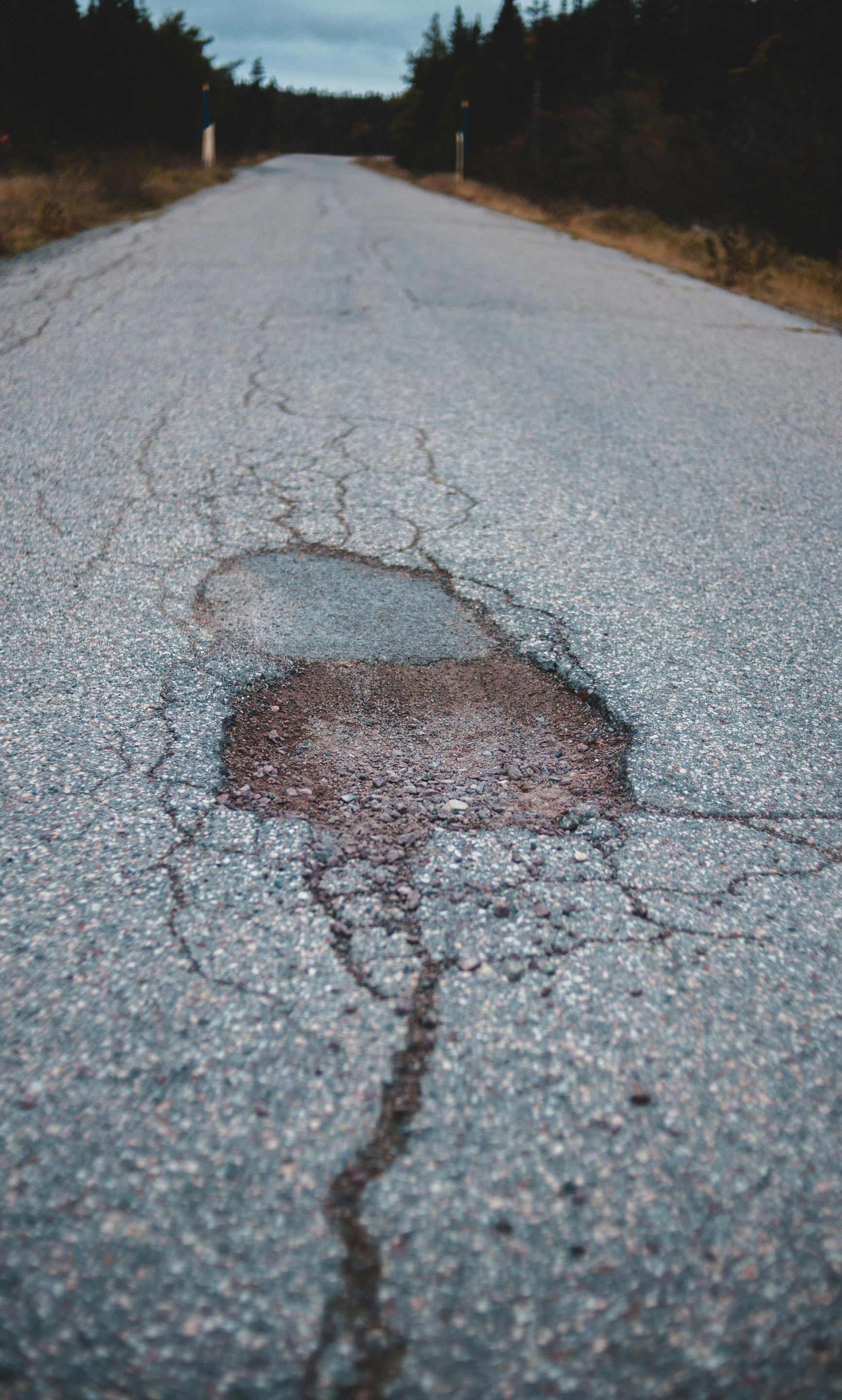 Cracked asphalt road with a large pothole, trees in the background, overcast sky.
