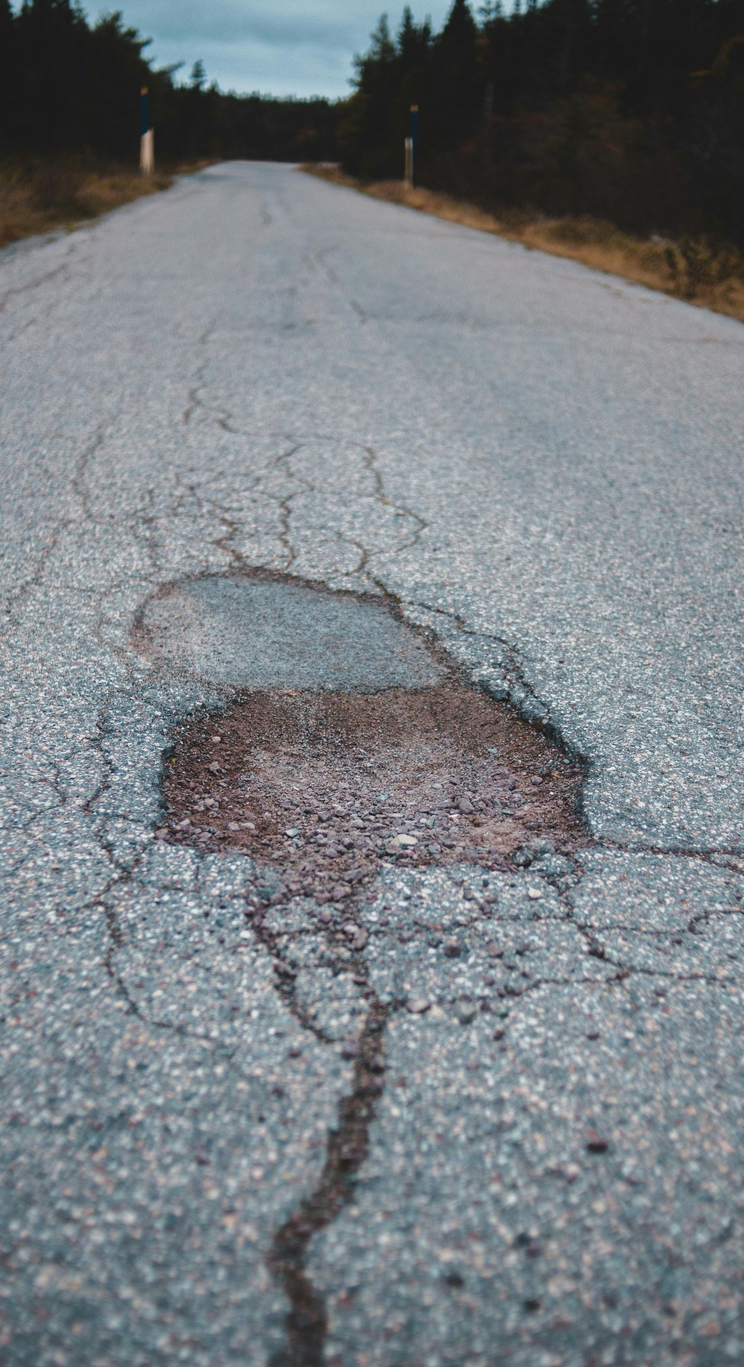 Cracked asphalt road with a large pothole, trees in the background, overcast sky.