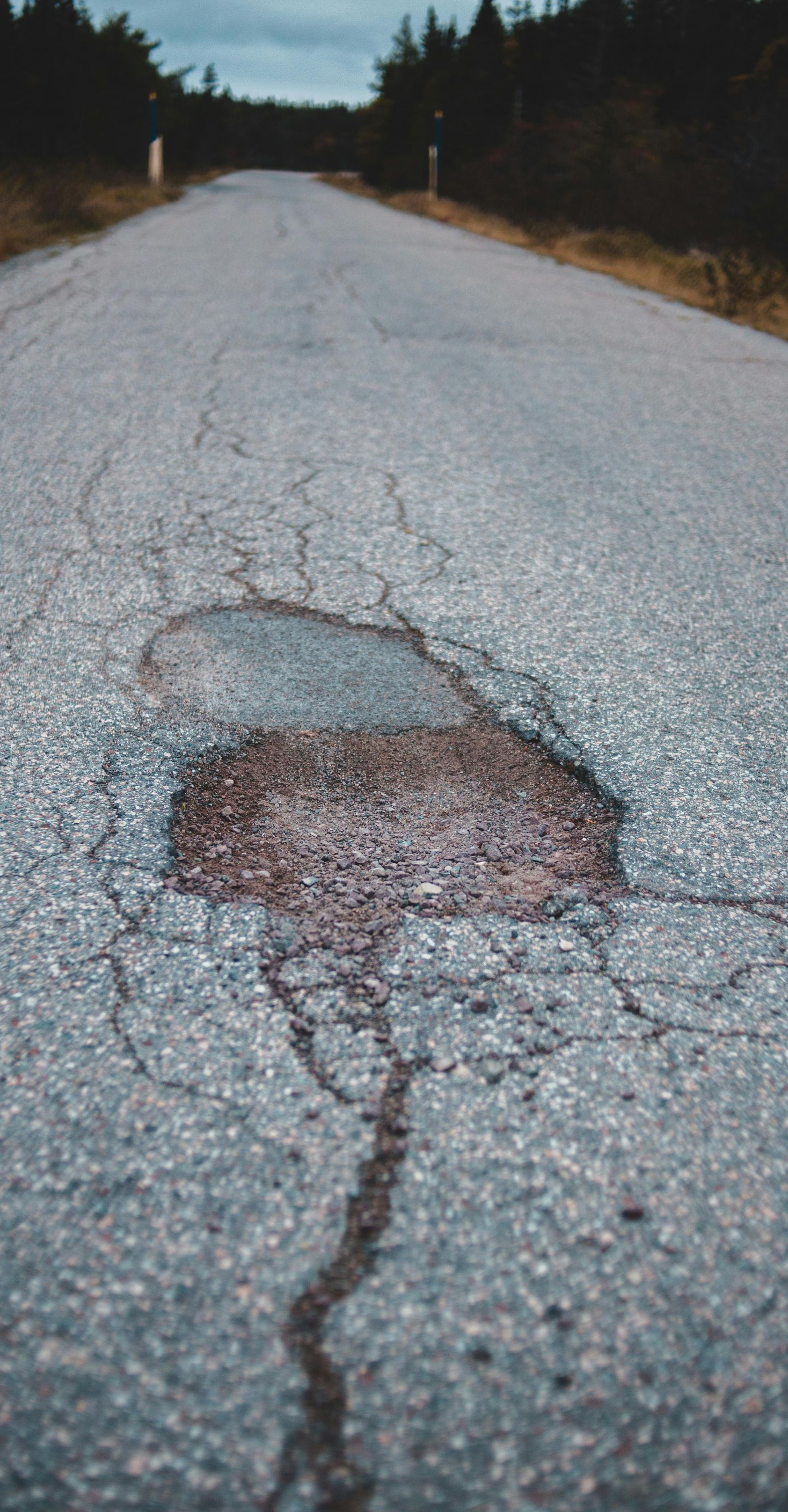 Cracked asphalt road with a large pothole, trees in the background, overcast sky.