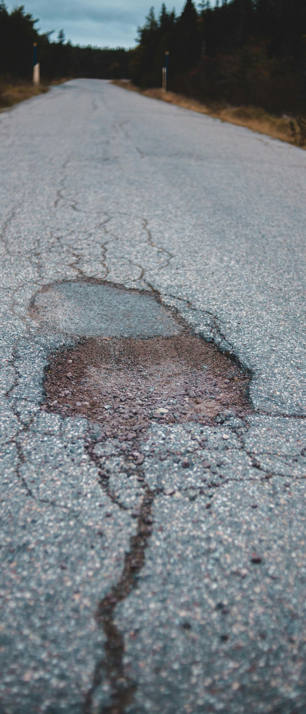 A pothole in the center of a weathered asphalt road stretching toward trees under an overcast sky.