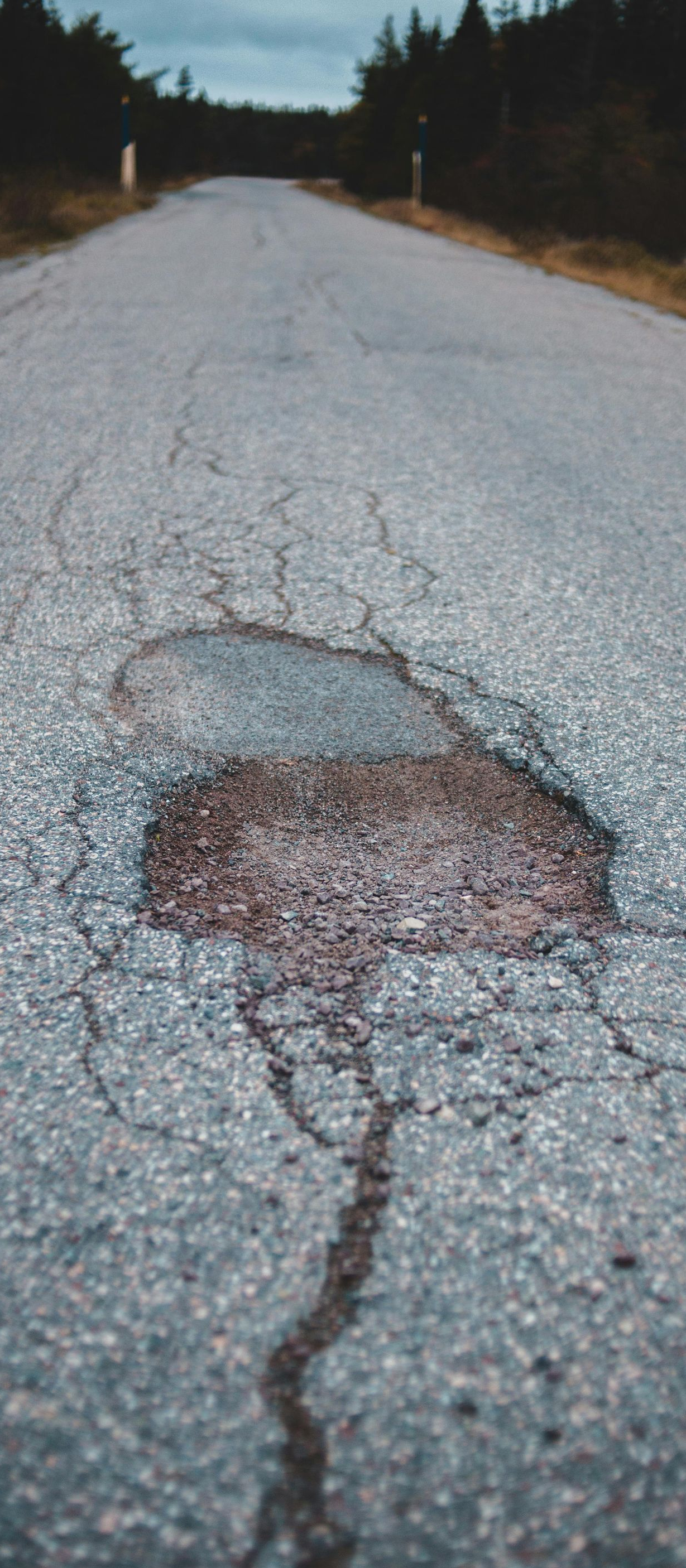 A pothole in the center of a weathered asphalt road stretching toward trees under an overcast sky.