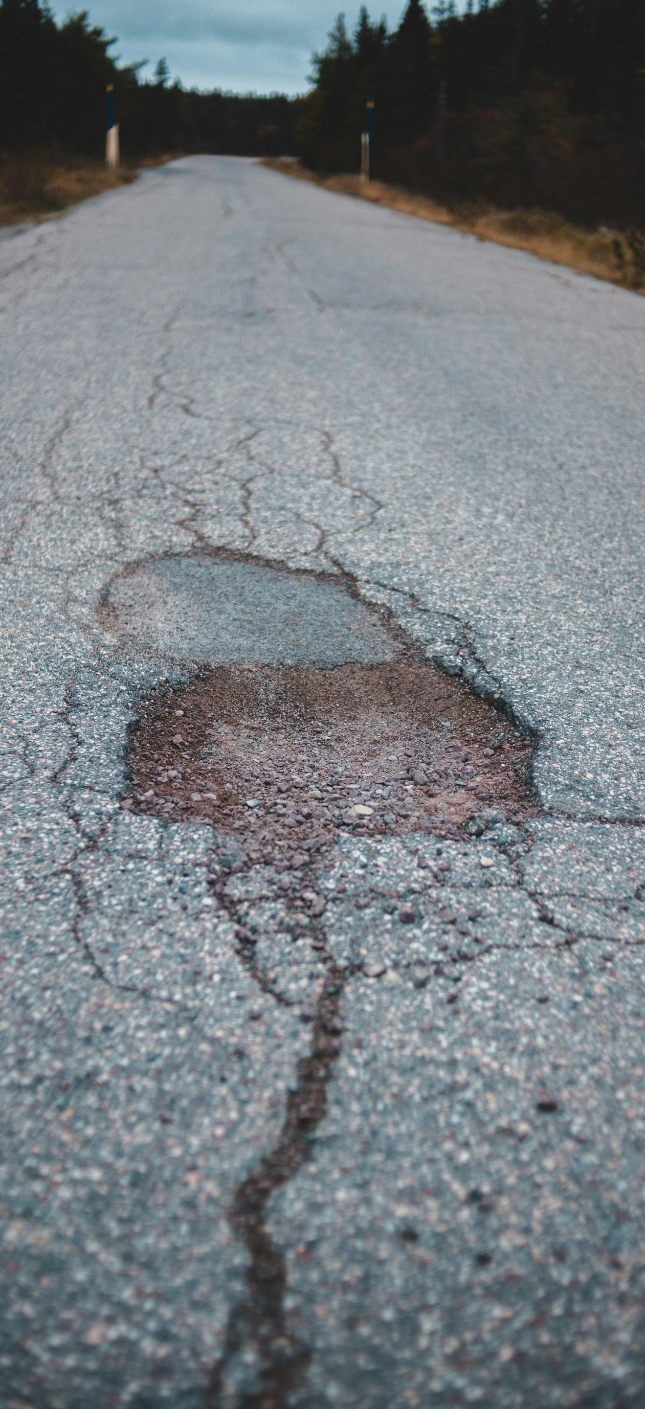 A pothole in the center of a weathered asphalt road stretching toward trees under an overcast sky.