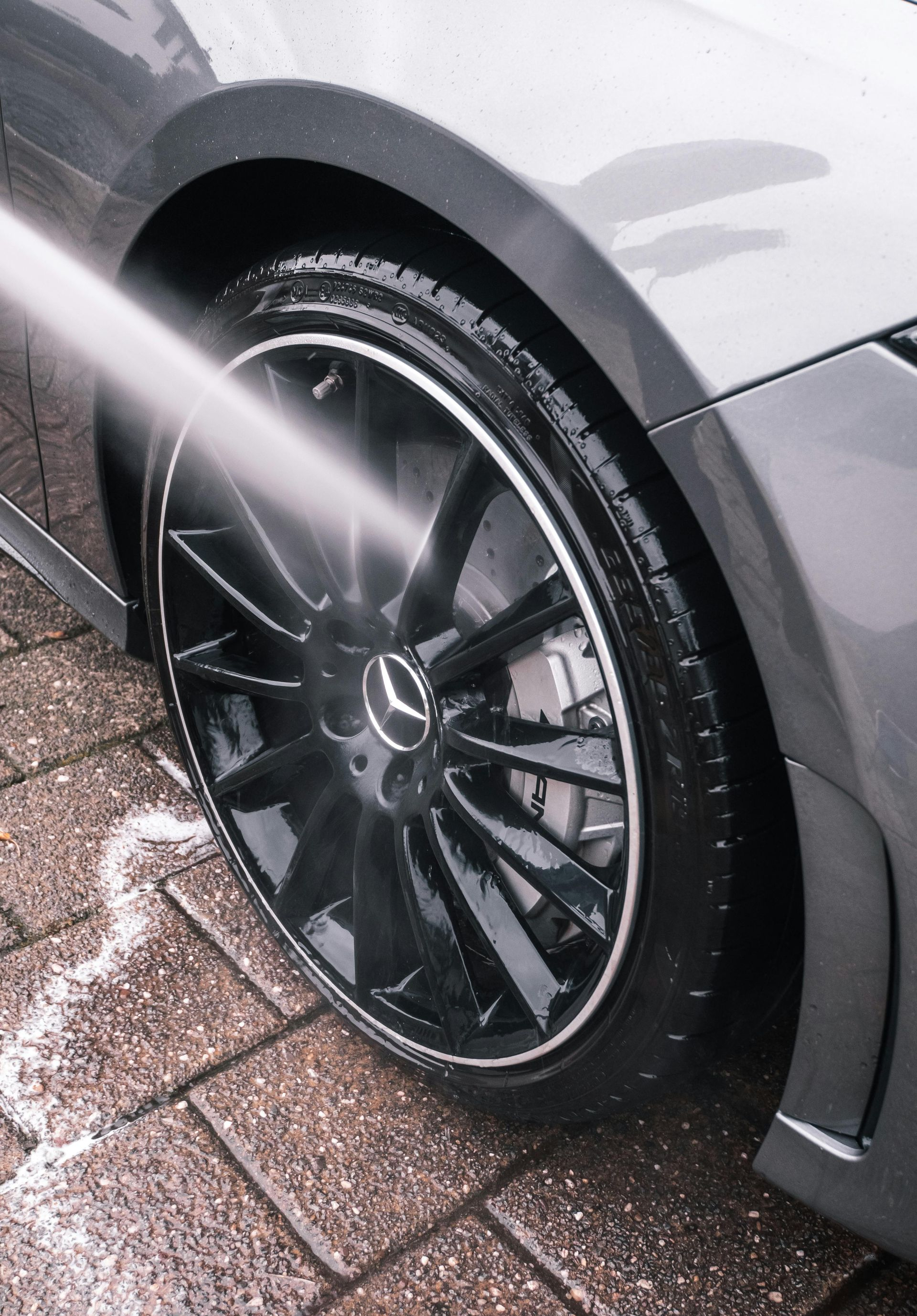 A pressure washer sprays water onto the shiny black wheel and tire of a gray Mercedes-Benz on a paved driveway.