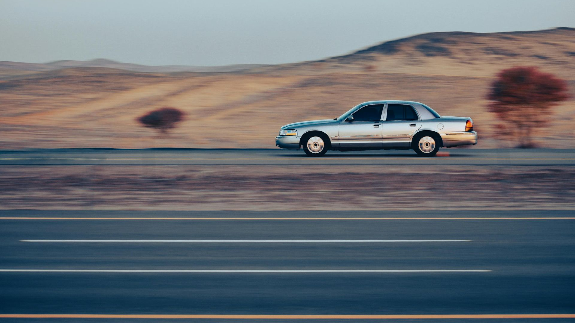 Silver sedan speeding on a desert highway, mountains and sparse trees in the background.