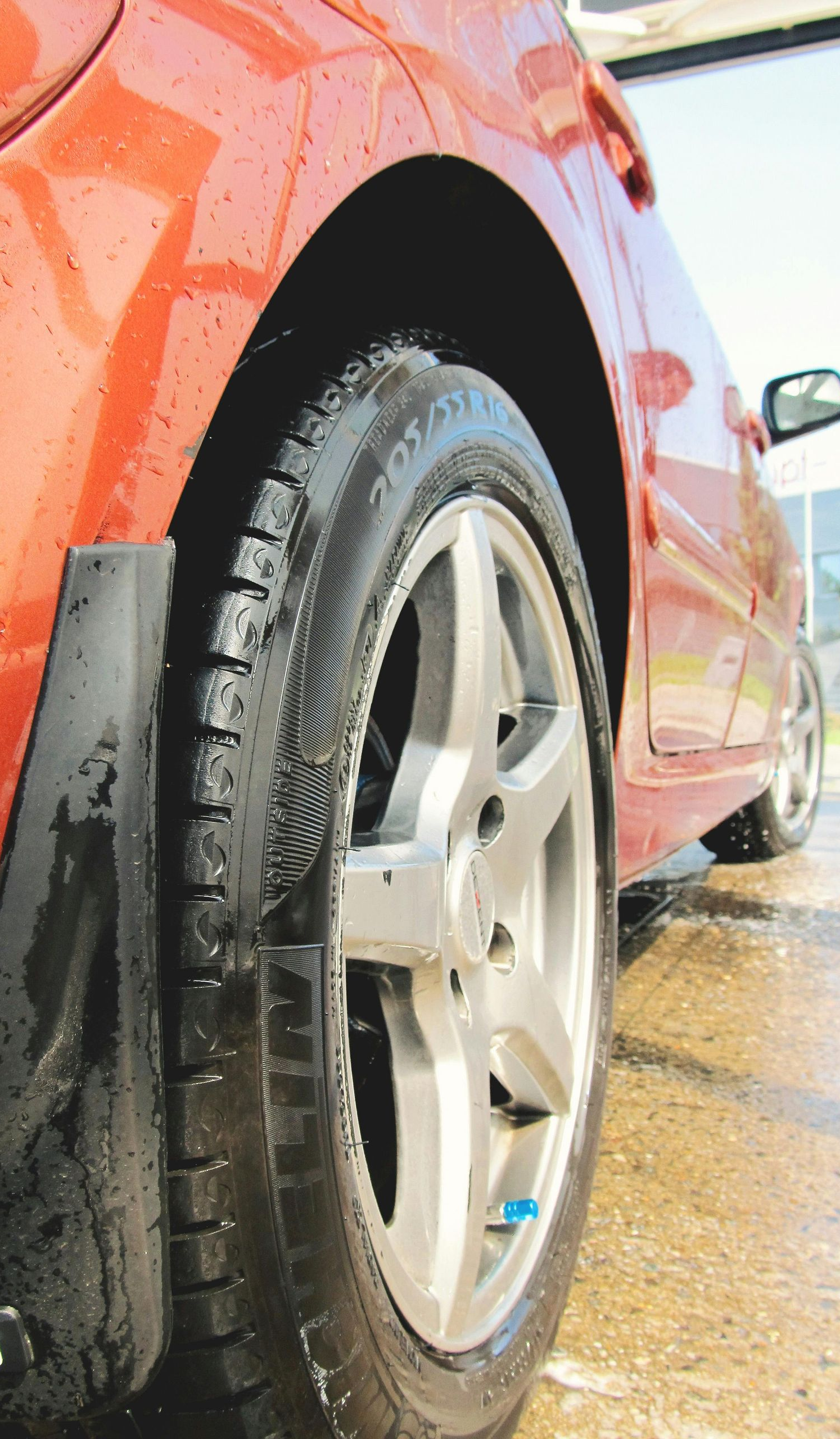 A close-up, low-angle view of a wet, orange car tire and silver rim on a paved surface during a wash.