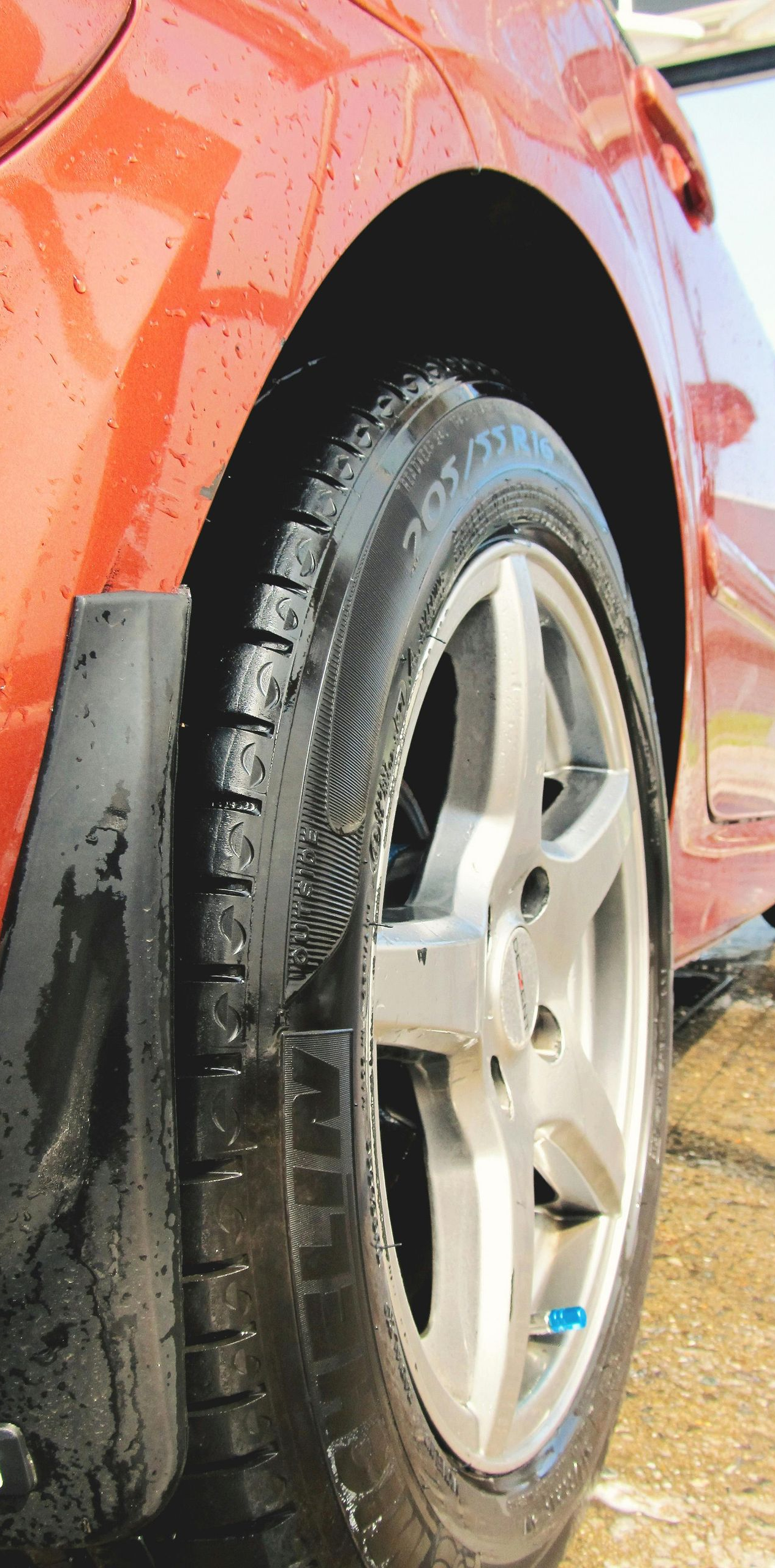A close-up, low-angle view of a wet, orange car tire and silver rim on a paved surface during a wash.