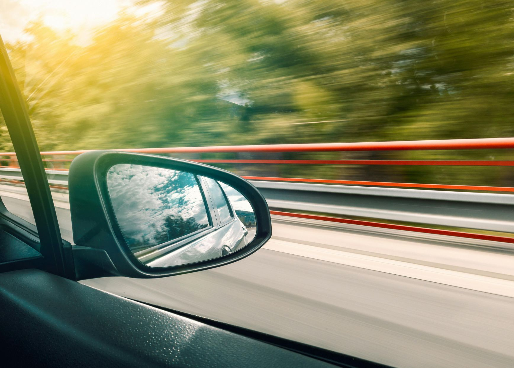 Car side mirror showing blurred scenery of road and trees, reflecting blue sky.