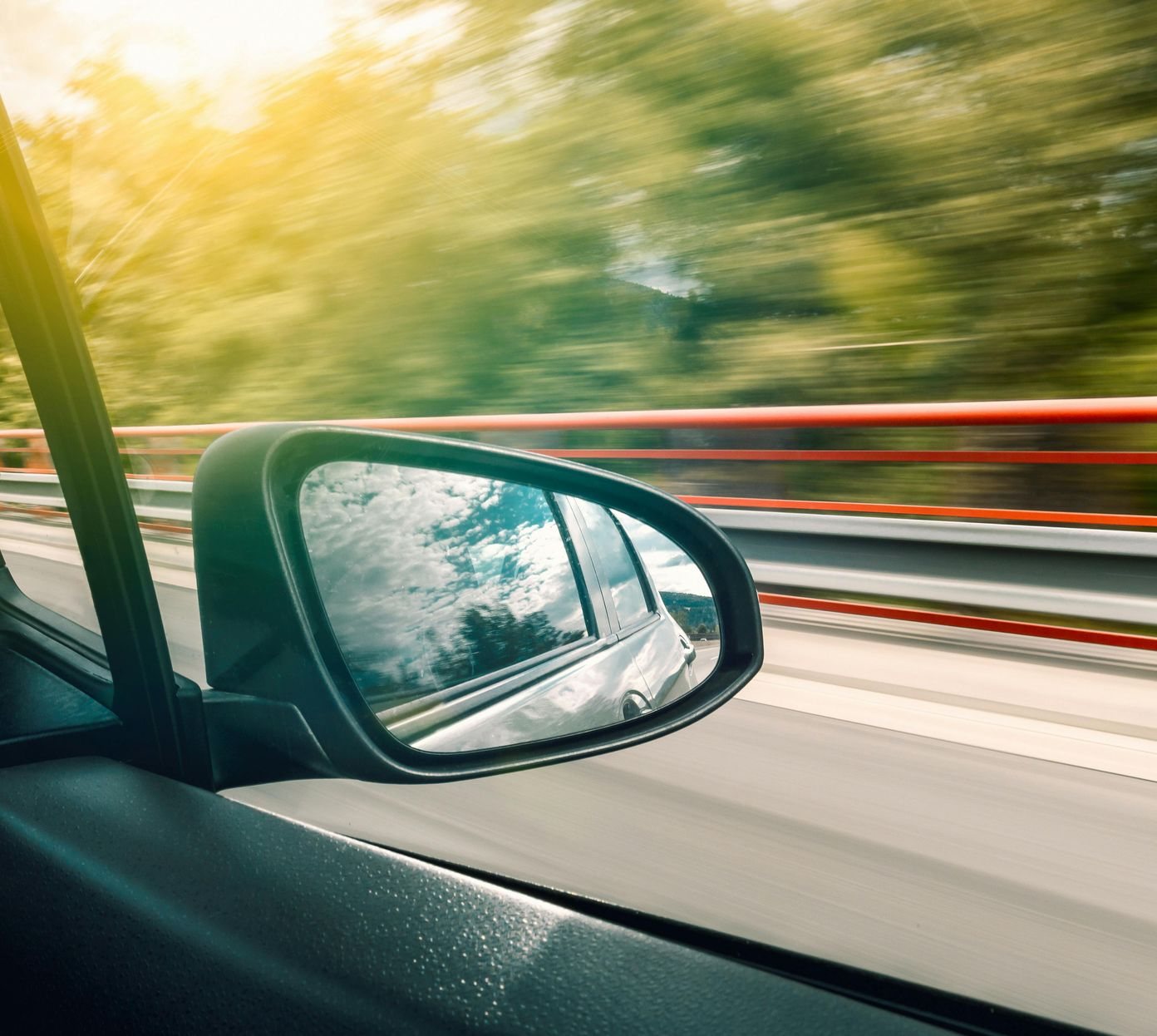 Car side mirror reflecting highway with blurred green trees; sunny day.