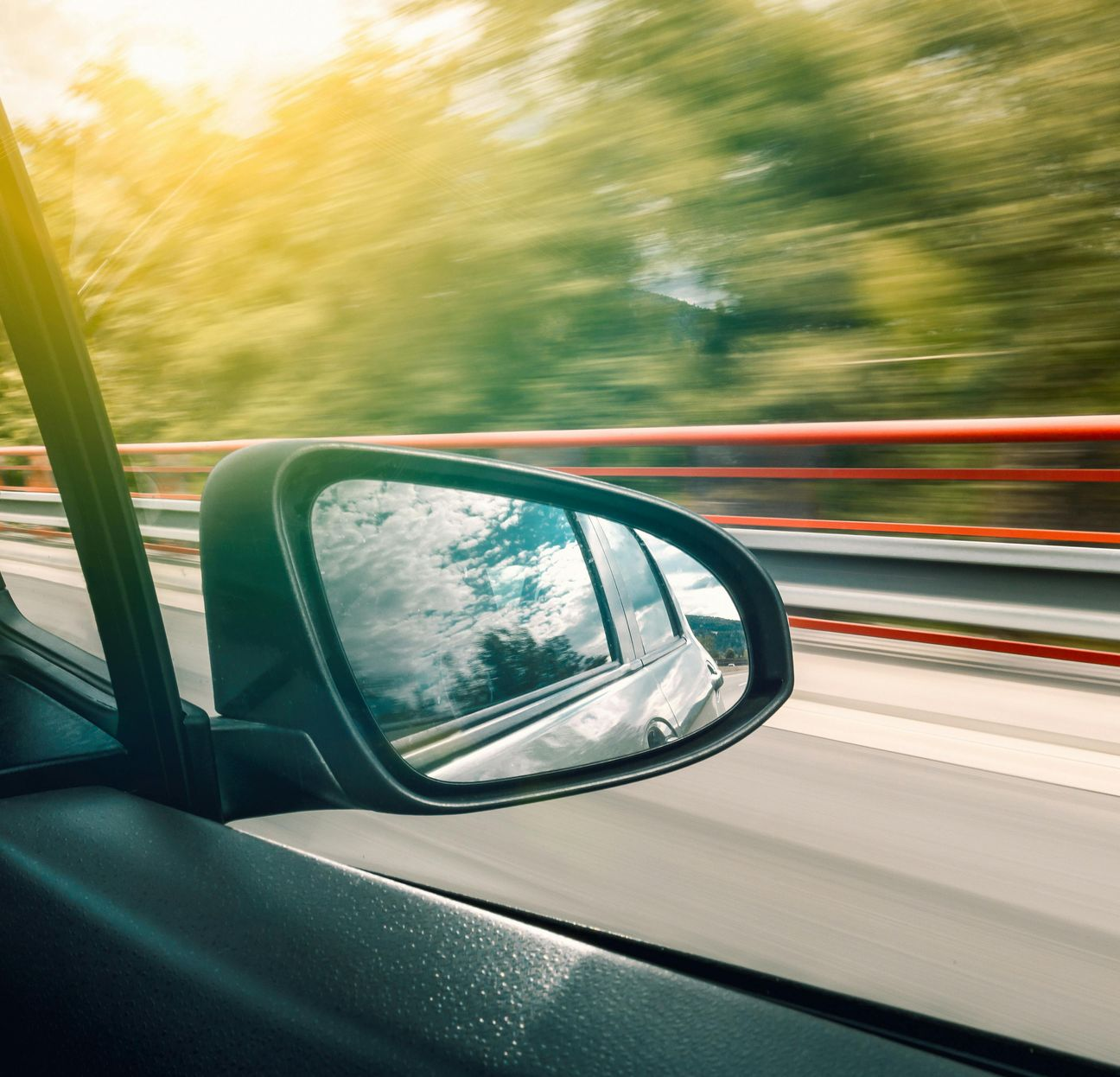 Car side mirror reflecting highway with blurred green trees; sunny day.