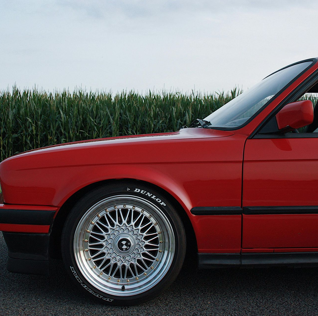 A red BMW E30 parked on a road next to a green cornfield.