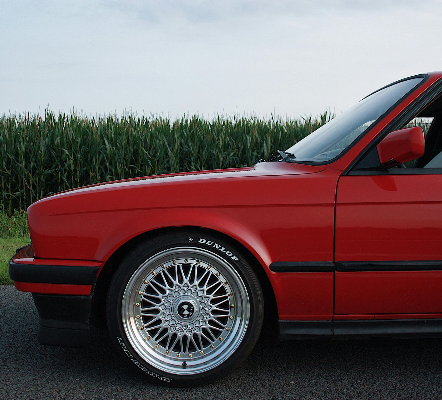 A red BMW E30 parked on a road next to a green cornfield.