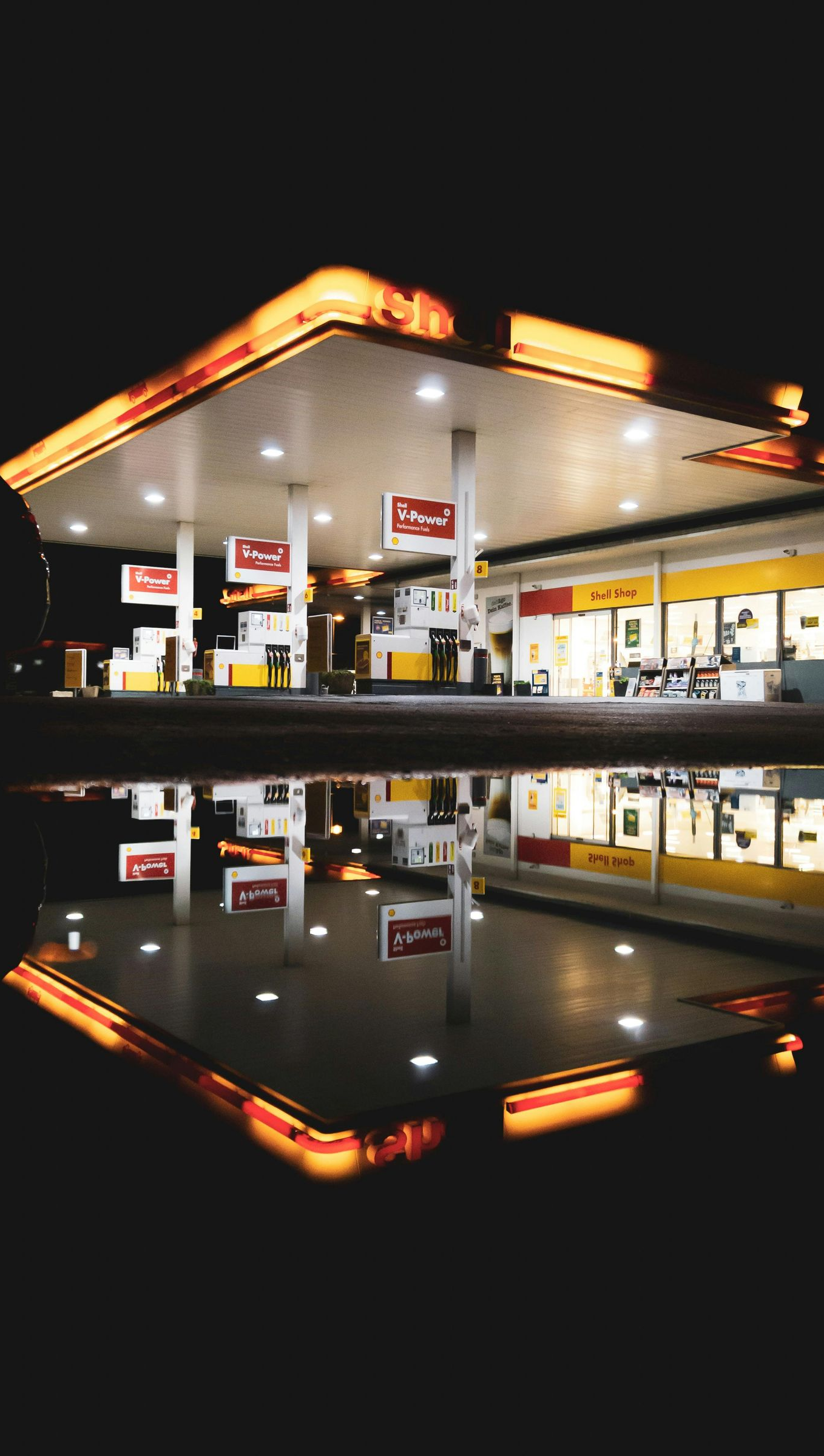 Gas station at night, illuminated with orange and white lights, reflected in a puddle.