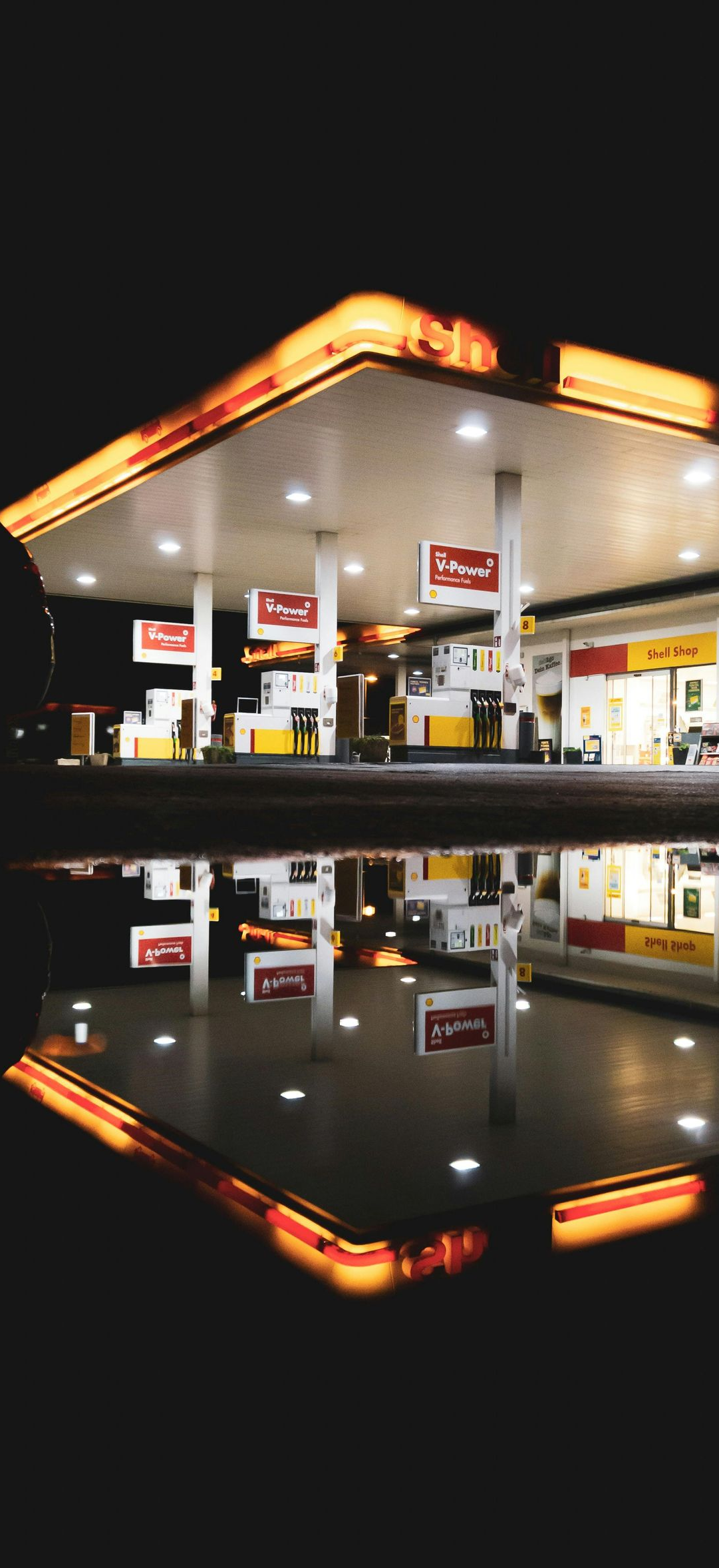 Gas station at night, illuminated with orange and white lights, reflected in a puddle.