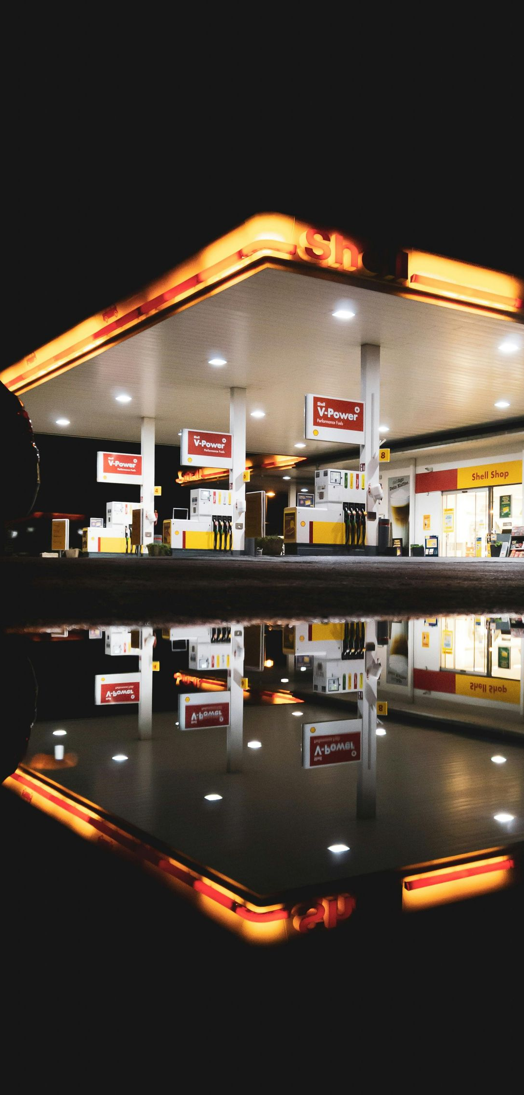 Gas station at night, illuminated with orange and white lights, reflected in a puddle.