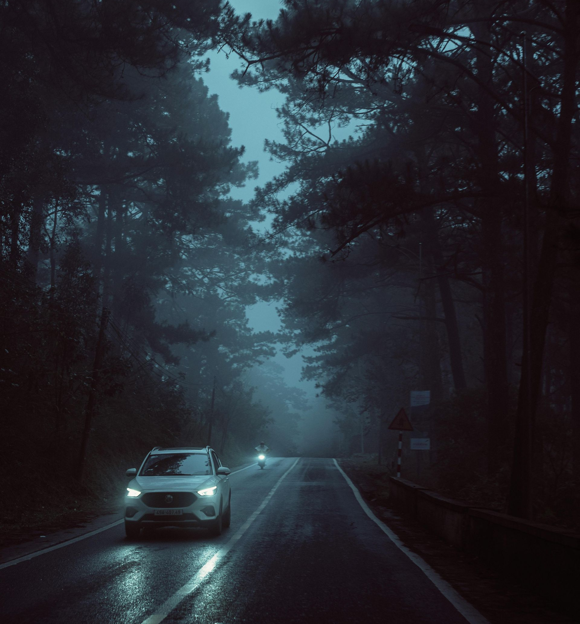 Car driving on a foggy, dark road through a forest at night. Headlights illuminate the wet asphalt.