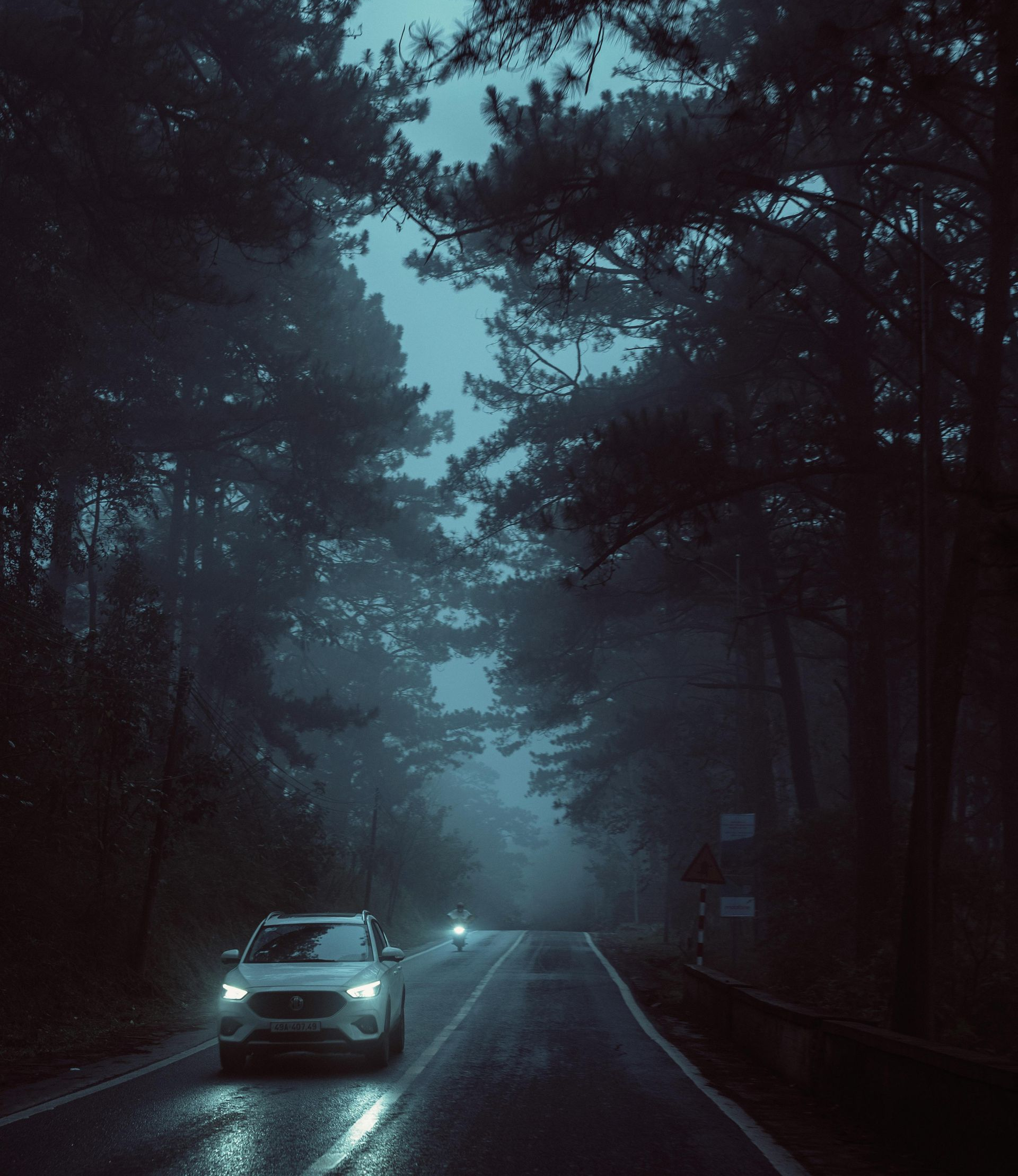 Car driving on a foggy, dark road through a forest at night. Headlights illuminate the wet asphalt.