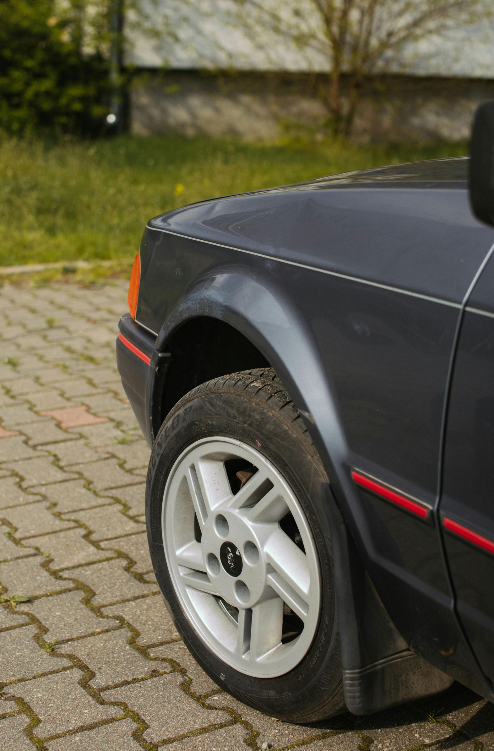 Gray car's front quarter, showing wheel, fender, and part of the hood. Red trim detail. Parked on paving stones.