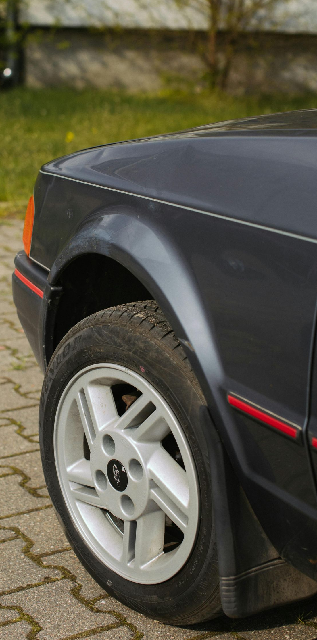 Dark grey Ford Escort front quarter panel, wheel on brick pavement.