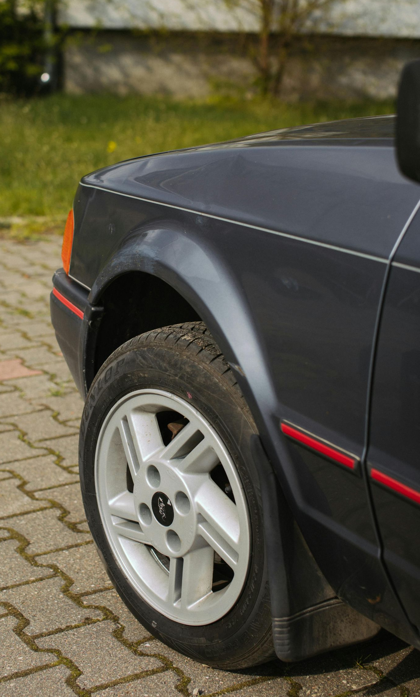 Dark grey Ford Escort front quarter panel, wheel on brick pavement.