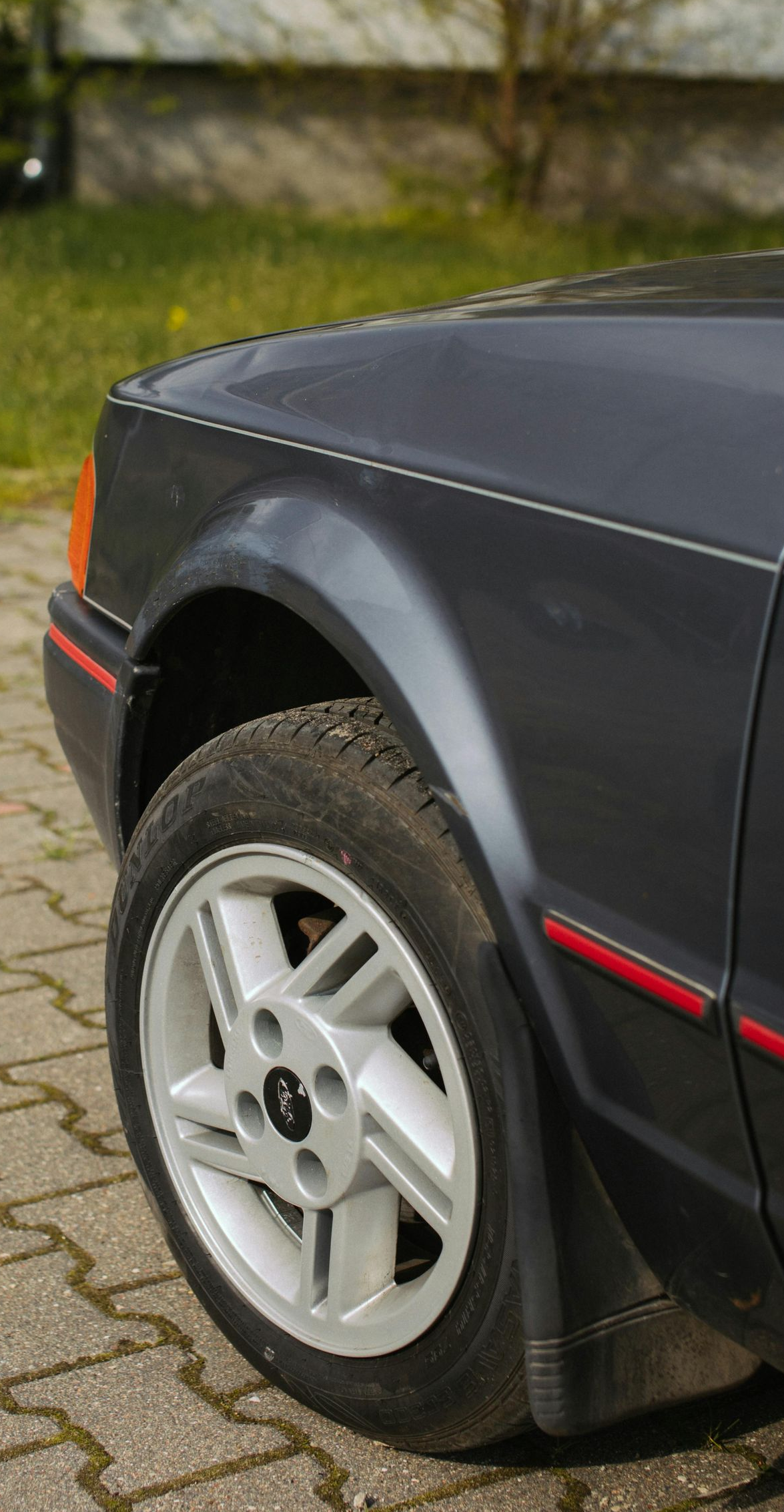 Dark grey Ford Escort front quarter panel, wheel on brick pavement.